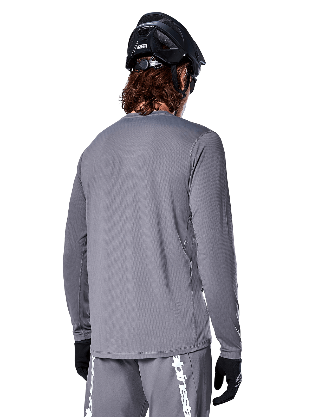 Jersey de manga larga Alpinestars A-Dura Rocker en gris oscuro, vista trasera de un ciclista de montaña, combinado con pantalones cortos grises a juego con logotipos en blanco, casco de MTB negro y guantes negros, equipamiento profesional de ciclismo todoterreno.