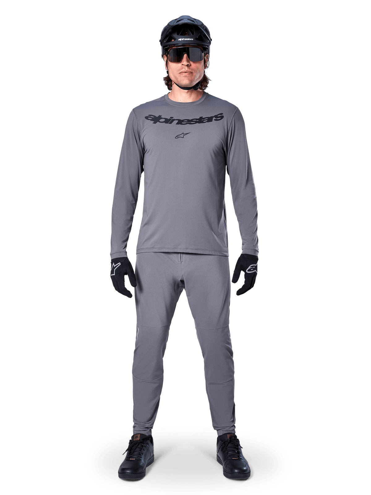 Ciclista masculino con el jersey de manga larga Alpinestars A-Dura Rocker en gris oscuro, pantalones grises a juego, casco de MTB negro, gafas de sol y guantes, equipamiento profesional de ciclismo de montaña para senderos todoterreno