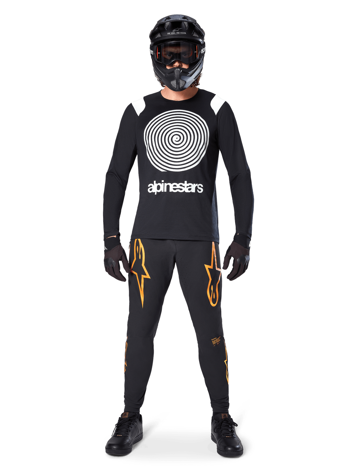 Modelo masculino con equipamiento completo para bicicleta de montaña, jersey de manga larga Alpinestars A-Supra Oscar en negro con gráfico de espiral blanco en el pecho, pantalones negros con logos de estrella naranjas, casco integral y gafas negras, guantes y zapatillas negras, atuendo coordinado para MTB de gravedad