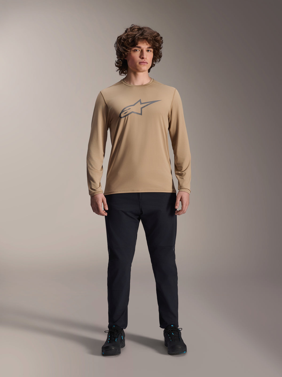 Alpinestars A-Dura Astar Jersey de manga larga, marrón claro con logotipo de Astars gris en el pecho, usado por un modelo con pantalones técnicos y zapatillas negros, equipamiento casual de alto rendimiento para ciclismo de montaña