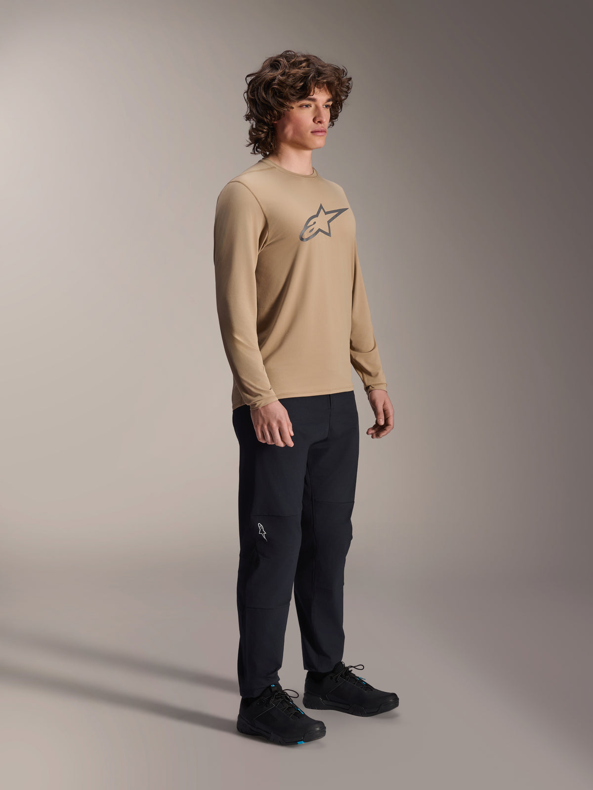 Alpinestars A-Dura Astar Long Sleeve Jersey, marrón claro, usado por un modelo con pantalones y calzado negros, con un logotipo Astars gris en el pecho, equipamiento de rendimiento casual para bicicleta de montaña