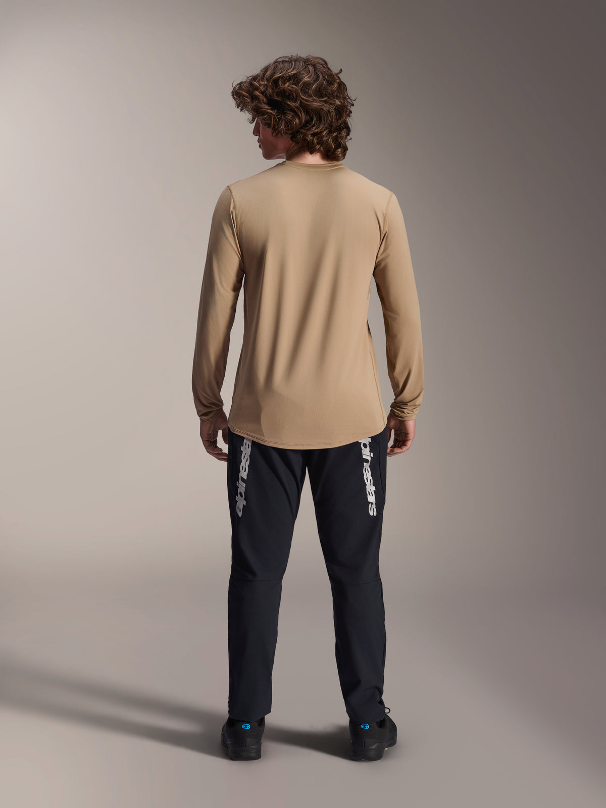 Alpinestars A-Dura Astar Long Sleeve Jersey, marrón claro, vista trasera que muestra la cola más larga, combinada con pantalones negros con el logotipo de Alpinestars, equipamiento de alto rendimiento para bicicleta de montaña