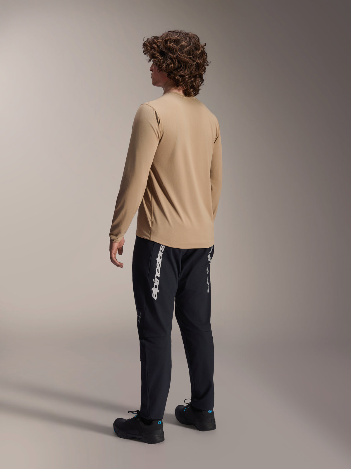 Jersey de manga larga Alpinestars A-Dura Astar en marrón claro, vista trasera de un modelo masculino que muestra el diseño de cola más larga, combinado con pantalones y calzado negros, equipamiento de rendimiento casual para bicicleta de montaña