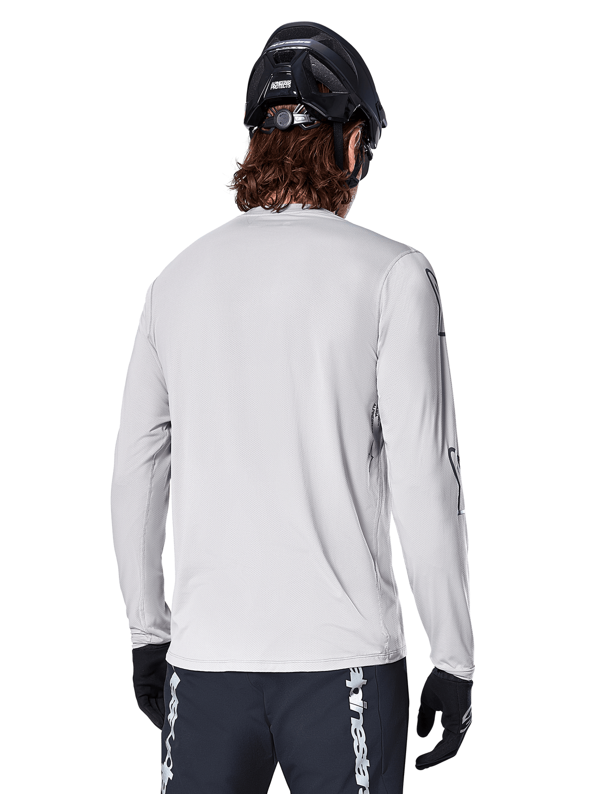 Jersey de manga larga Alpinestars A-Dura Switch, Gris Claro, vista trasera de un ciclista de montaña con casco de MTB negro y guantes, con una parte trasera más larga para mayor cobertura y tejido jacquard texturizado, equipamiento de ciclismo de alto rendimiento.
