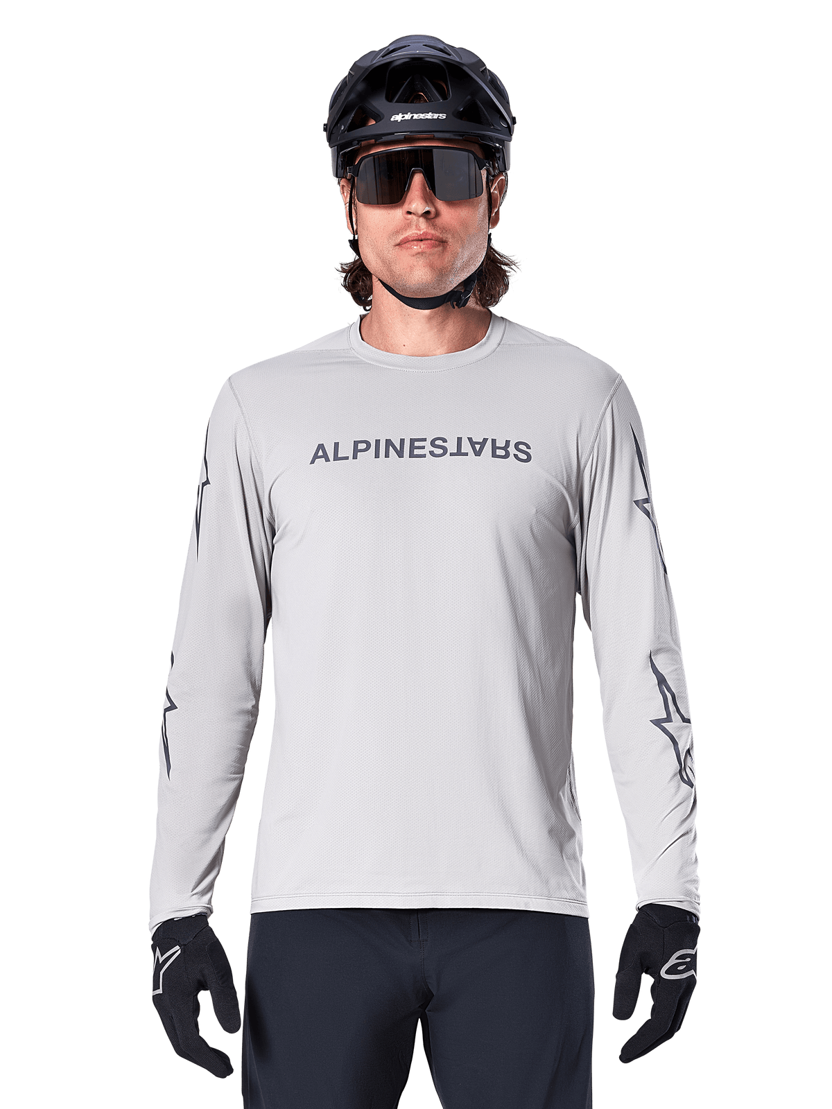 Alpinestars A-Dura Switch Long Sleeve Jersey, gris claro, presenta el nombre de la marca en gris oscuro en el pecho y logotipos de estrellas en las mangas, usado por un ciclista con casco de MTB y guantes negros, ropa de ciclismo de alto rendimiento para trail