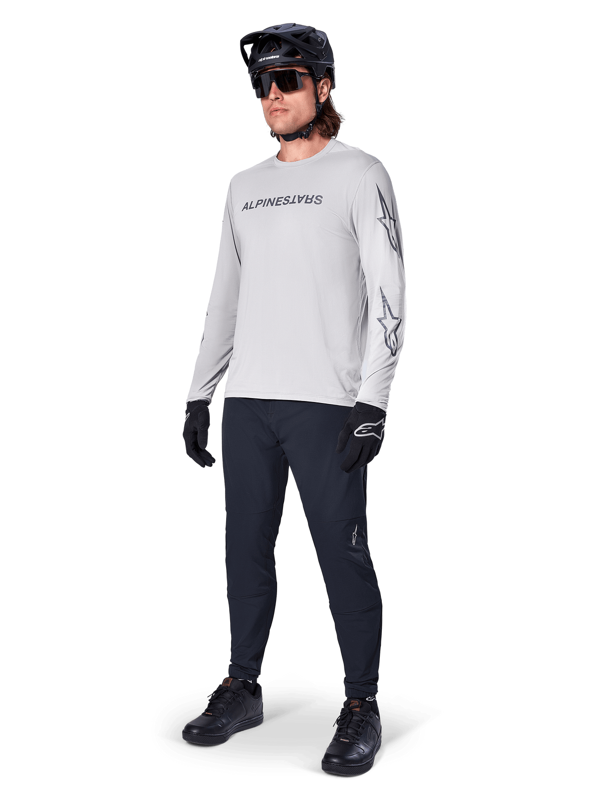 Jersey de manga larga Alpinestars A-Dura Switch, gris claro, usado por un ciclista con pantalones negros de montaña y casco, con logotipo de la marca en el pecho y gráficos de estrellas en las mangas, equipamiento de alto rendimiento para ciclismo todoterreno