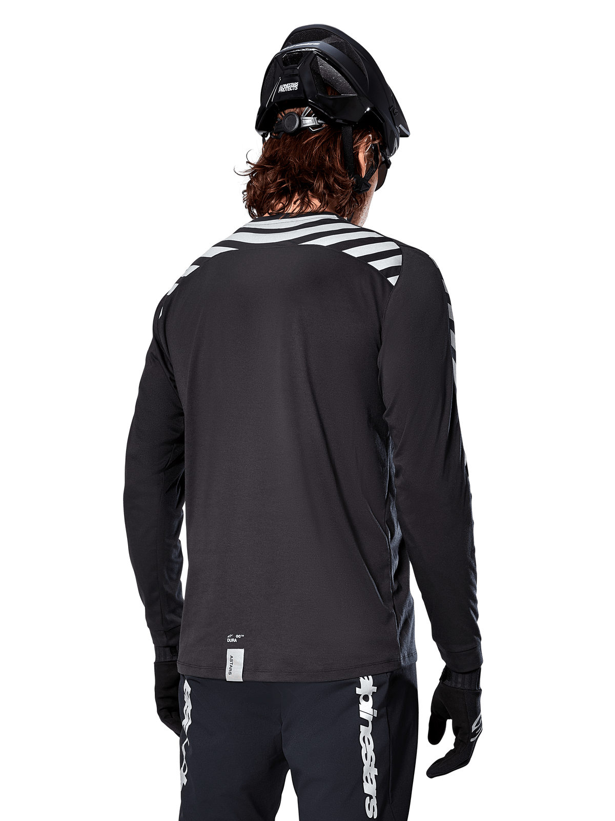 Jersey de manga larga Alpinestars A-Dura Dri Oscar, negro reflectante, vista trasera que muestra franjas diagonales reflectantes en los hombros y una parte trasera más larga, usado con casco y guantes de MTB negros, equipamiento técnico para ciclismo de montaña