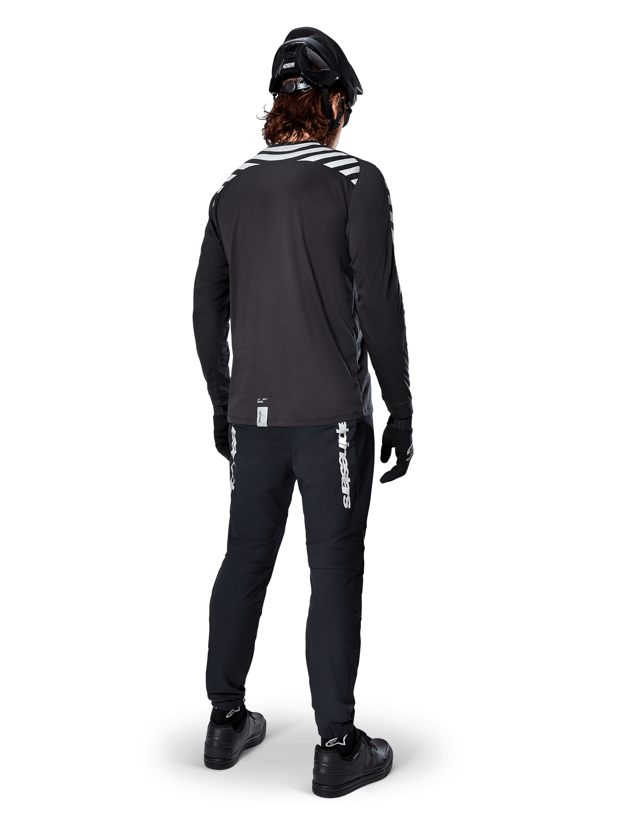 Mountain bike rider wearing the Alpinestars A-Dura Dri Oscar Long Sleeve Jersey in Black Reflective, viewed from behind, paired with black technical pants, helmet, and gloves, featuring reflective stripe graphics on the shoulders and a longer drop tail for off-road cycling.