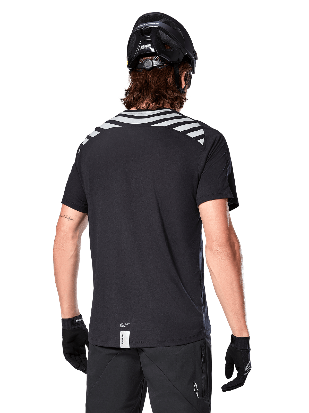 Jersey Alpinestars A-Dura Dri Oscar, jersey de MTB de manga corta, negro reflectante, vista trasera de un modelo con casco de ciclismo y guantes, con rayas grises reflectantes en los hombros y parte trasera caída, tejido técnico transpirable para ciclismo de montaña