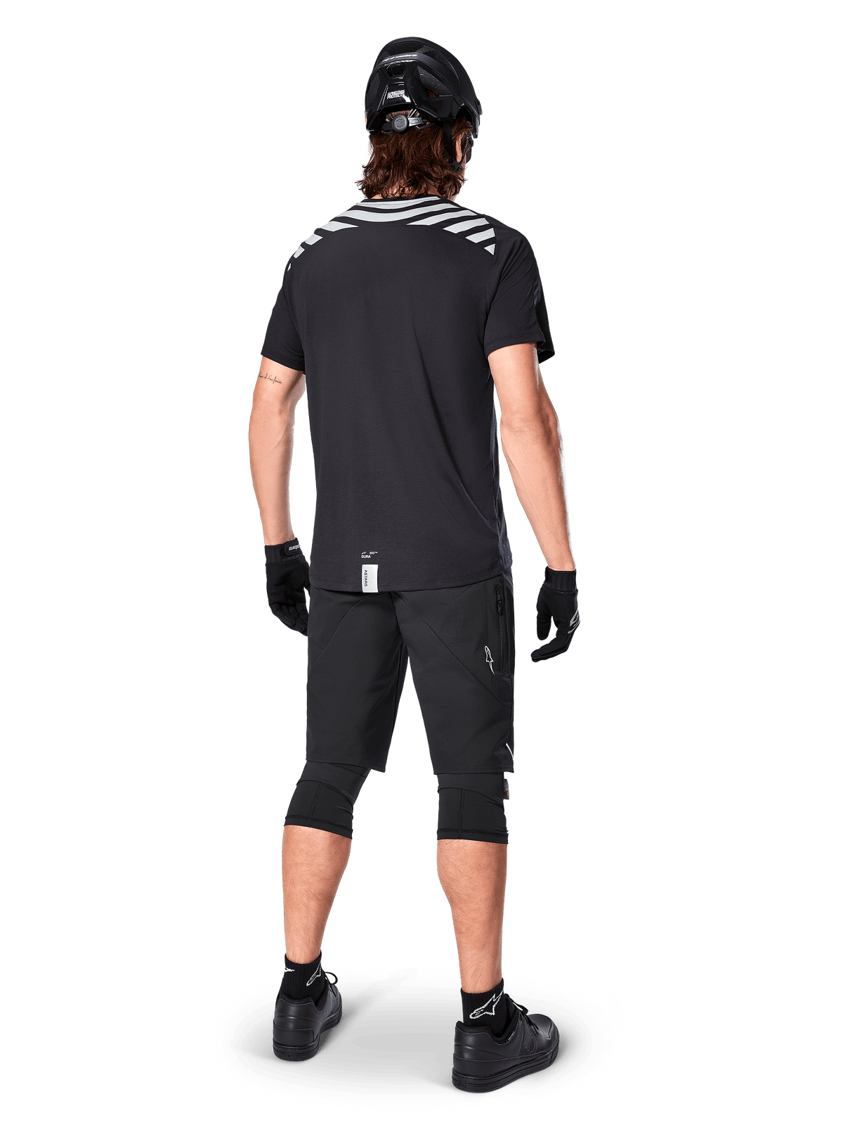 Jersey de manga corta Alpinestars A-Dura Dri Oscar, negro reflectante, vista trasera de un ciclista usando el jersey con pantalones cortos de montaña negros, casco y rodilleras, con gráficos reflectantes en los hombros y diseño de cola caída para MTB.