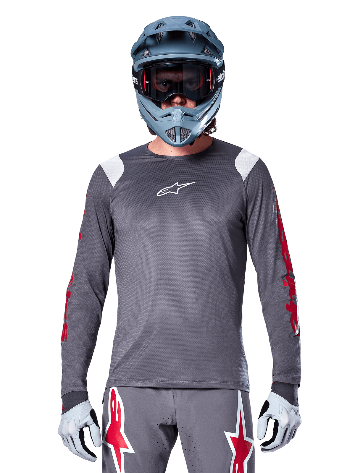 MTB rider wearing Alpinestars A-Supra Astar Long Sleeve Jersey, Dark Gray with red graphics on sleeves and white shoulder accents, matching gray pants with red star logos, blue full-face helmet and goggles, coordinated off-road cycling gear