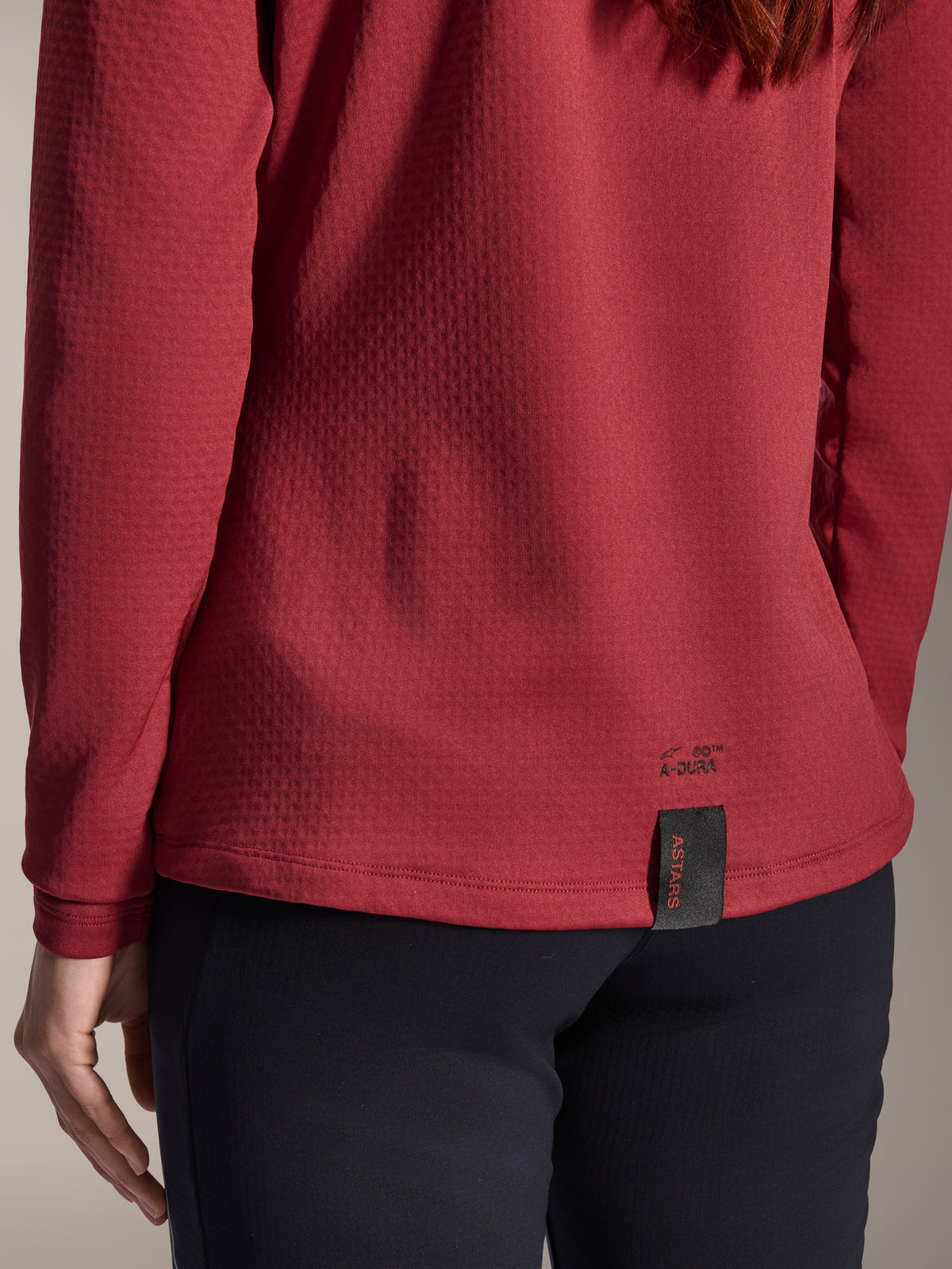 Jersey Térmico Alpinestars Stella A-Dura para mujer, color burdeos, vista de cerca del dobladillo trasero con la marca A-Dura y etiqueta tejida ASTARS, tejido de vellón aislante texturizado, ropa de bicicleta de montaña para mujer