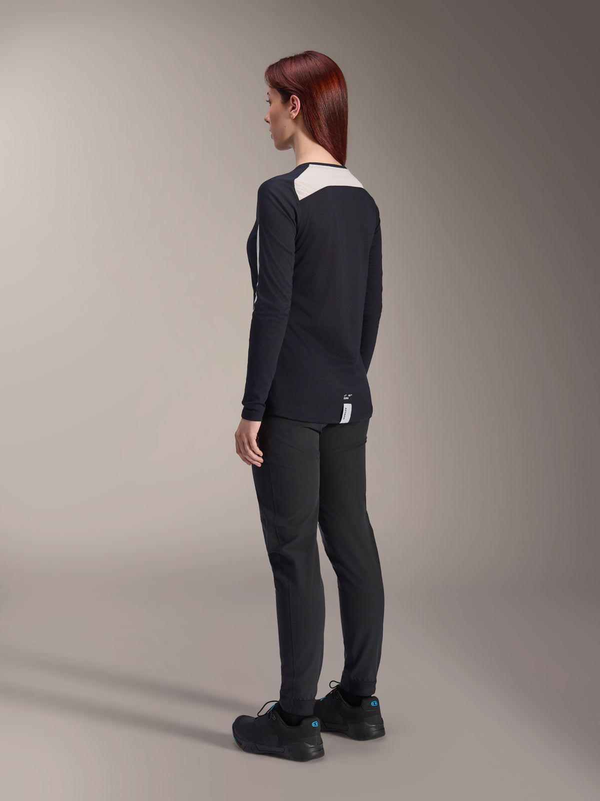 Mujer ciclista vistiendo pantalones Stella A-Dura y jersey Stella A-Dura Elite de Alpinestars para mujer, negro, vista posterior que muestra paneles de malla blancos en los hombros del jersey de manga larga, pantalones técnicos de corte ajustado, equipamiento coordinado de alto rendimiento para bicicleta de montaña.