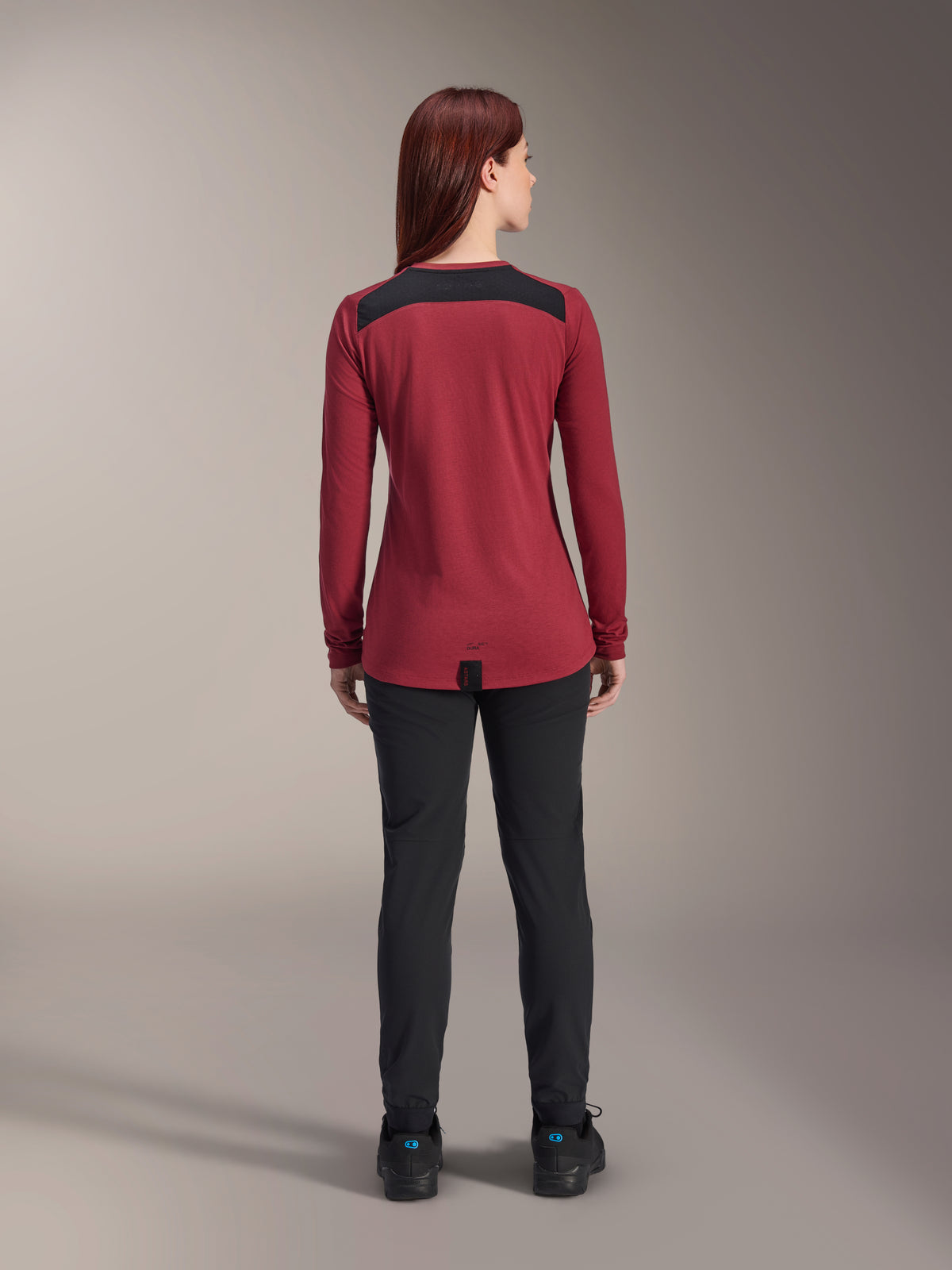 Jersey Alpinestars Stella A-Dura Dri Astar de manga larga para mujer, rojo borgoña con paneles de malla negros en los hombros, ropa técnica de MTB vista desde atrás con pantalones negros, diseño de cola caída para mayor cobertura al pedalear