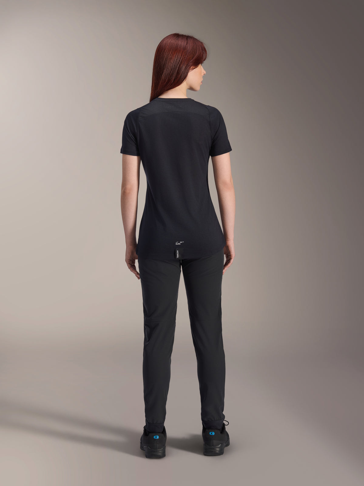 Alpinestars Jersey Stella A-Dura Dri Switch de manga corta para mujer, negro, vista trasera en una modelo, con cola más larga y paneles de malla técnica, combinado con pantalones negros, equipamiento de alto rendimiento para ciclismo MTB