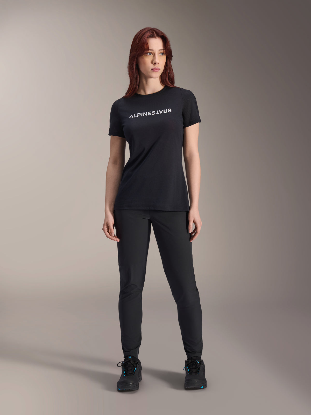 Modelo luciendo el jersey de manga corta Alpinestars Stella A-Dura Dri Switch para mujer en color negro, con logotipo blanco en el pecho, combinado con pantalones técnicos negros y calzado deportivo negro, fotografía de estudio de cuerpo completo
