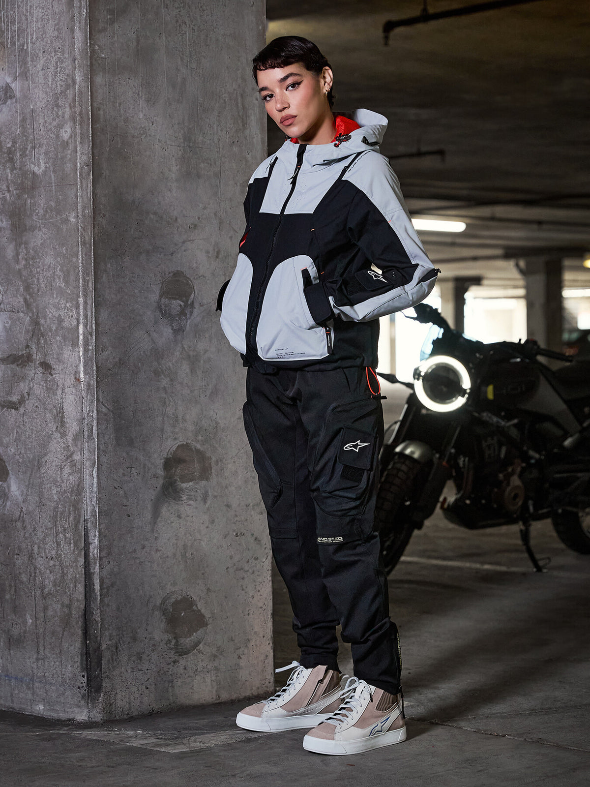 Alpinestars Actea Women's Pants, Black, female rider wearing cargo-style motorcycle pants with a grey and black technical jacket and beige sneakers, urban parking garage setting with motorcycle.