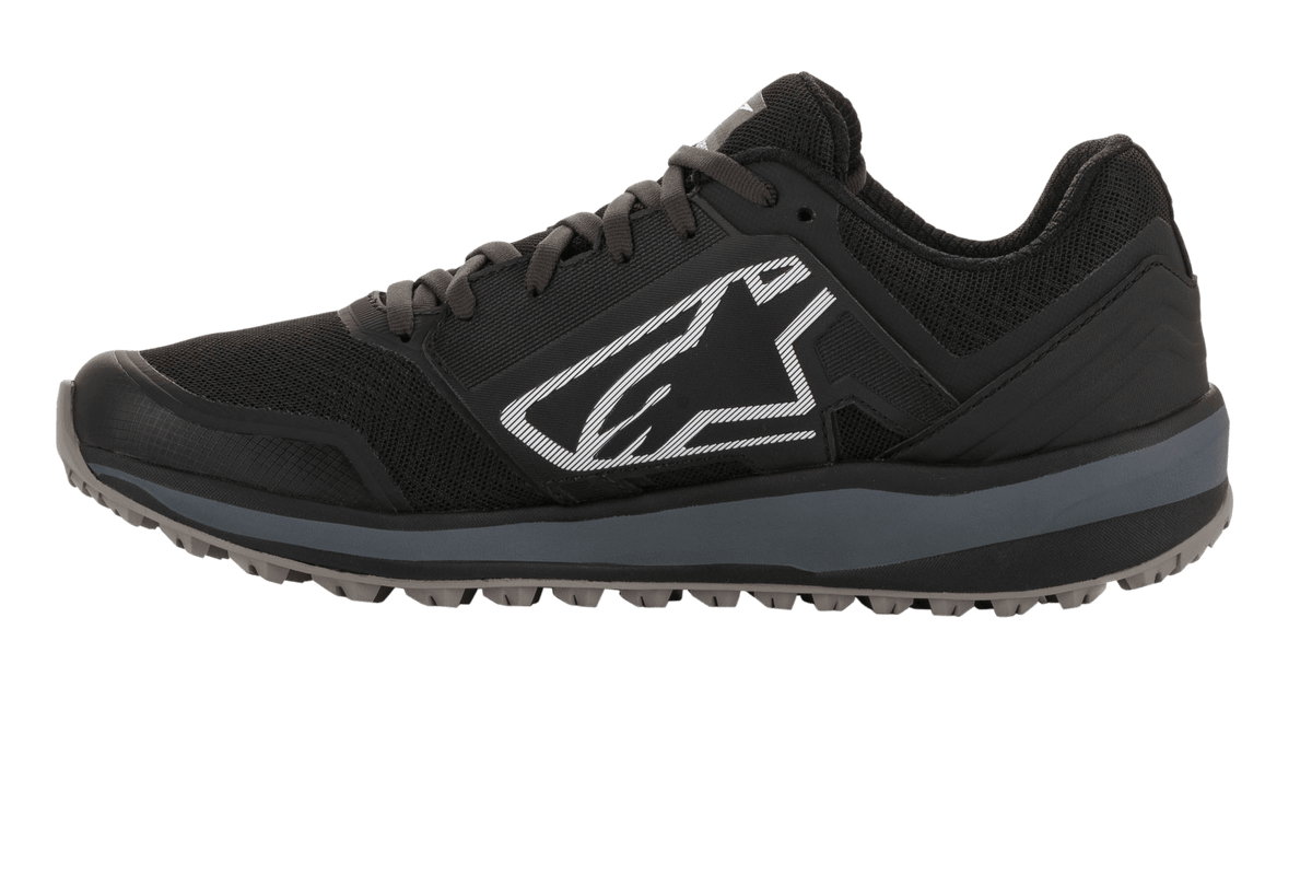 Alpinestars Meta Trail Shoes, calzado de running de alto rendimiento, negro y gris oscuro, perfil lateral con gran logotipo Astars blanco, entresuela gris oscuro, suela de goma con tacos resistente al aceite, diseñado para comodidad en el paddock y uso por equipos de automovilismo