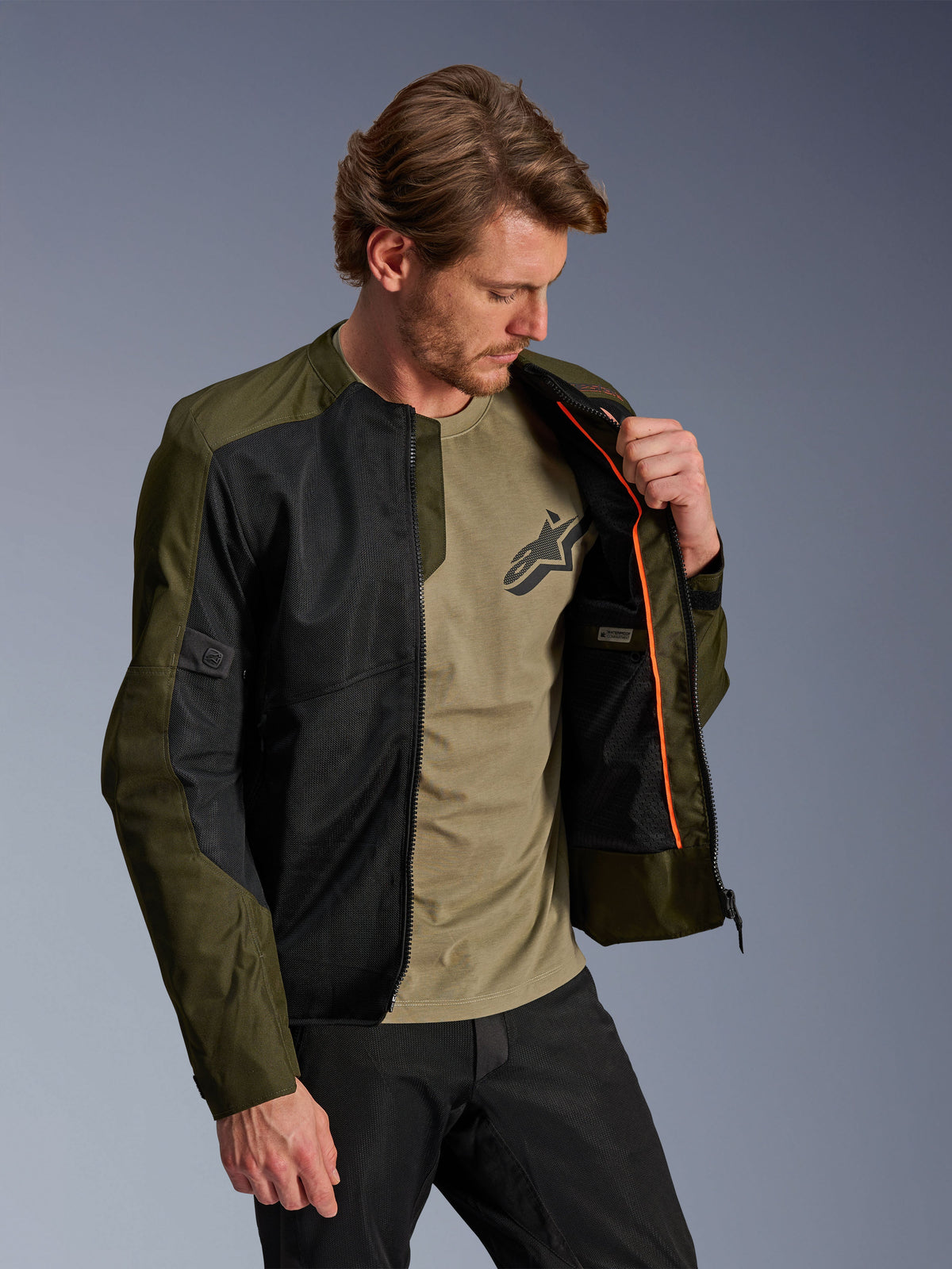 Chaqueta Alpinestars C-1 Air en caqui y negro, usada por un modelo masculino para mostrar el forro interior naranja y el compartimento impermeable, con grandes paneles de malla técnica para el flujo de aire, de estilo urbano contemporáneo combinada con una camiseta oliva a juego y pantalones negros