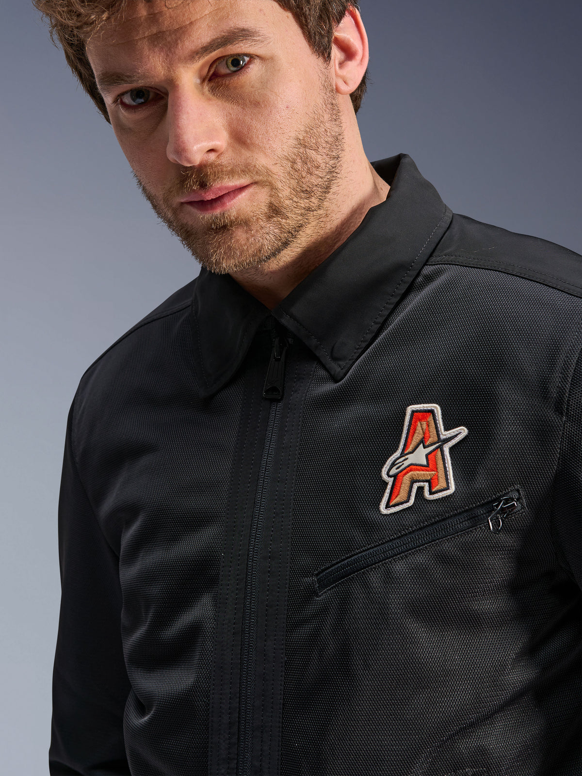 Chaqueta Alpinestars Ignite Air, negra, con un cuello de camisa tradicional y construcción de malla transpirable, destacada por un parche con el logotipo 'A' en rojo y dorado en el pecho, usada por un modelo masculino, ropa urbana para motociclista