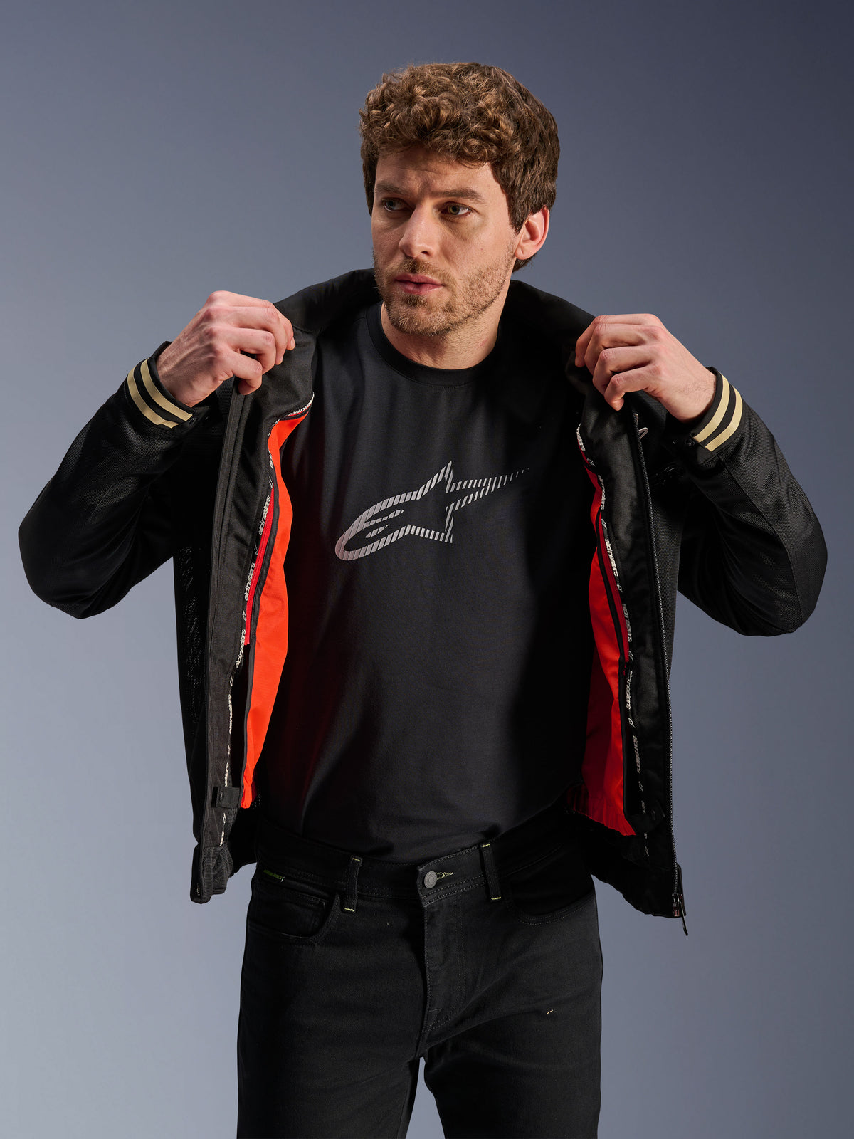 Hombre vistiendo la chaqueta Alpinestars Ignite Air negra, con cuerpo de malla y forro cortaviento desmontable en naranja brillante, combinada con una camiseta negra con logo de Astars y pantalones negros, estilo de motociclismo urbano contemporáneo