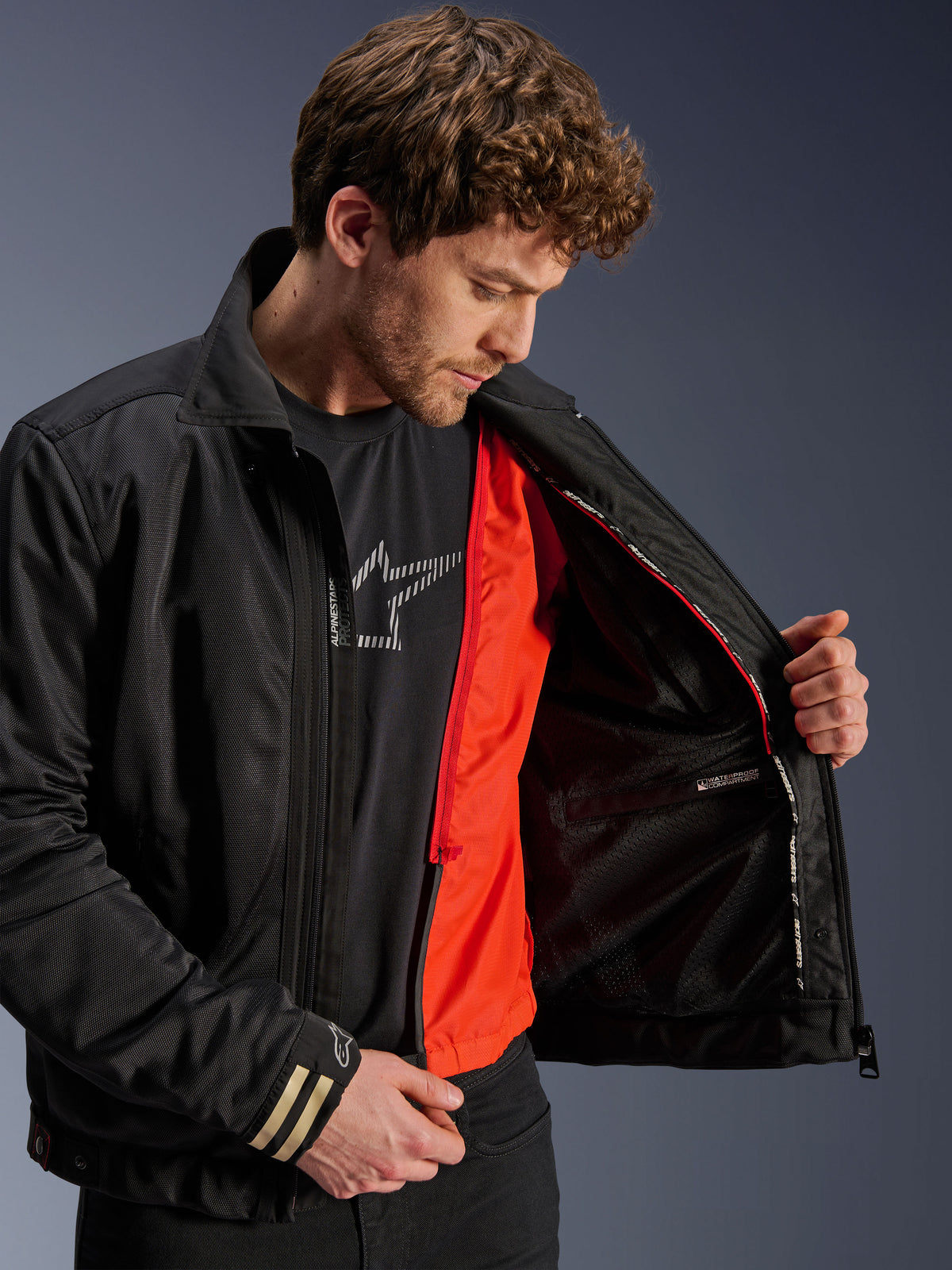 Chaqueta Alpinestars Ignite Air, chaqueta de moto urbana negra, con un forro cortaviento desmontable de color naranja brillante y cuerpo de malla, vista interior que muestra el compartimento impermeable, usada con una camiseta negra con el logo de Alpinestars