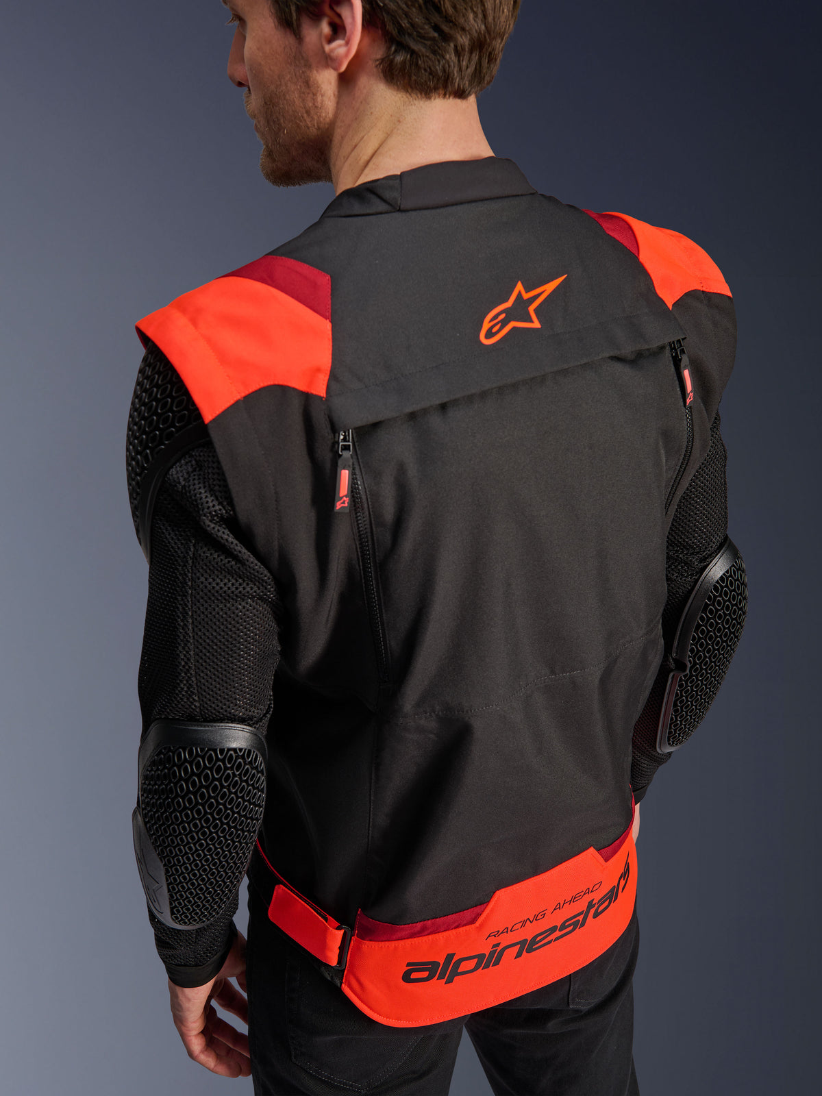 Chaqueta Alpinestars T-Stunt Air, negro, rojo flúor y rojo oscuro, vista posterior en un modelo, que muestra mangas de malla con protectores de codo con textura de panal, el logotipo de la estrella de Alpinestars en la parte superior de la espalda y una marca llamativa en la parte inferior con el texto 'RACING AHEAD', diseño modular de corte deportivo para motociclismo.