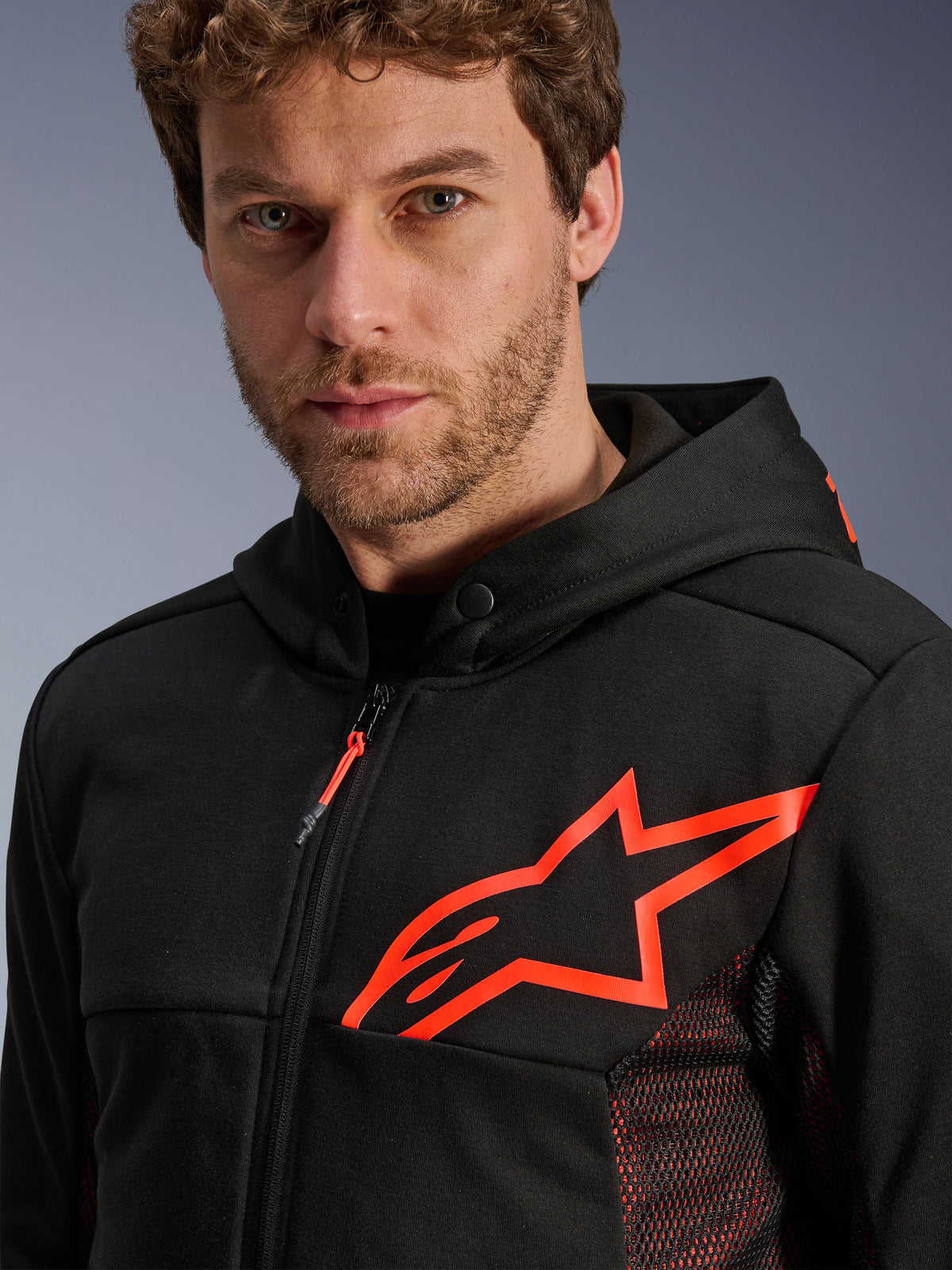 Alpinestars Chrome Air Sport Hoodie, negro y rojo flúor, primer plano que muestra el gran logotipo de estrella en rojo flúor en el pecho, paneles laterales de malla roja para ventilación, frontal con cremallera y cuello con botón de presión, equipamiento casual de moto para conducción urbana