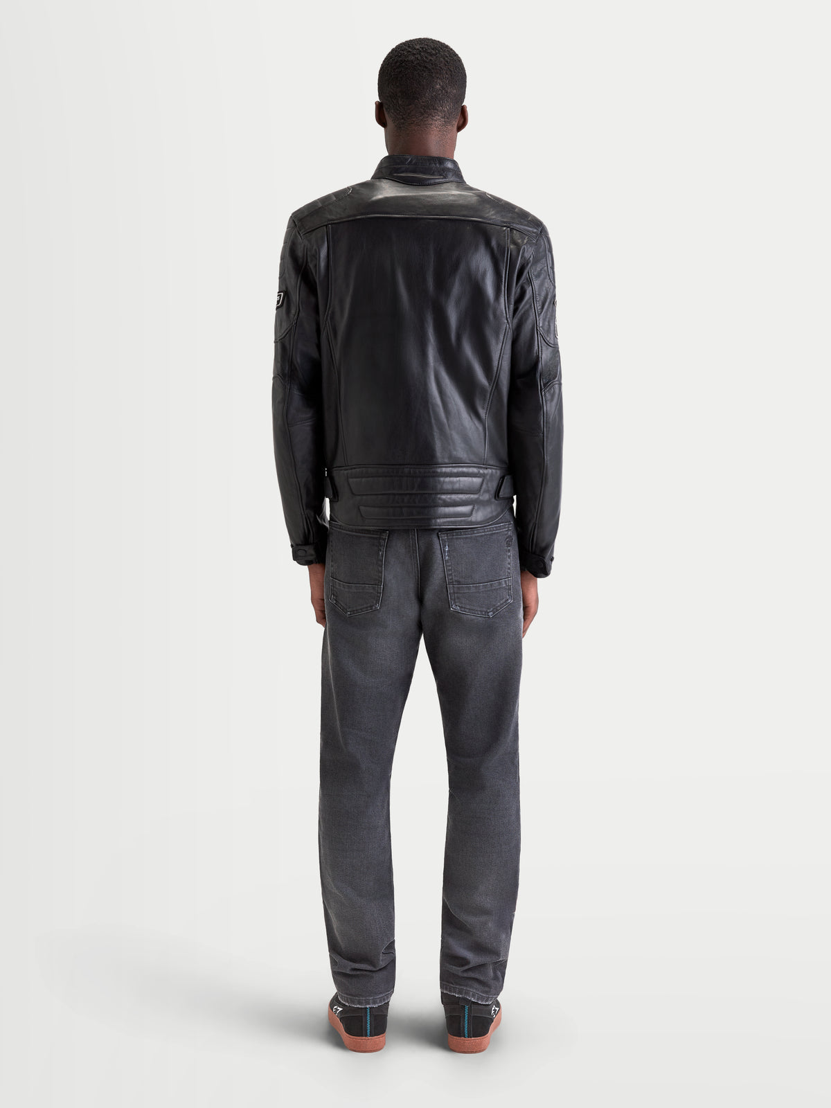 Alpinestars Revern chaqueta de cuero, chaqueta de moto urbana contemporánea para hombre en color negro, vista trasera en un modelo que lleva vaqueros y zapatos de moto casuales, mostrando la construcción de cuero bovino flor envejecido y el estilo de motociclista clásico