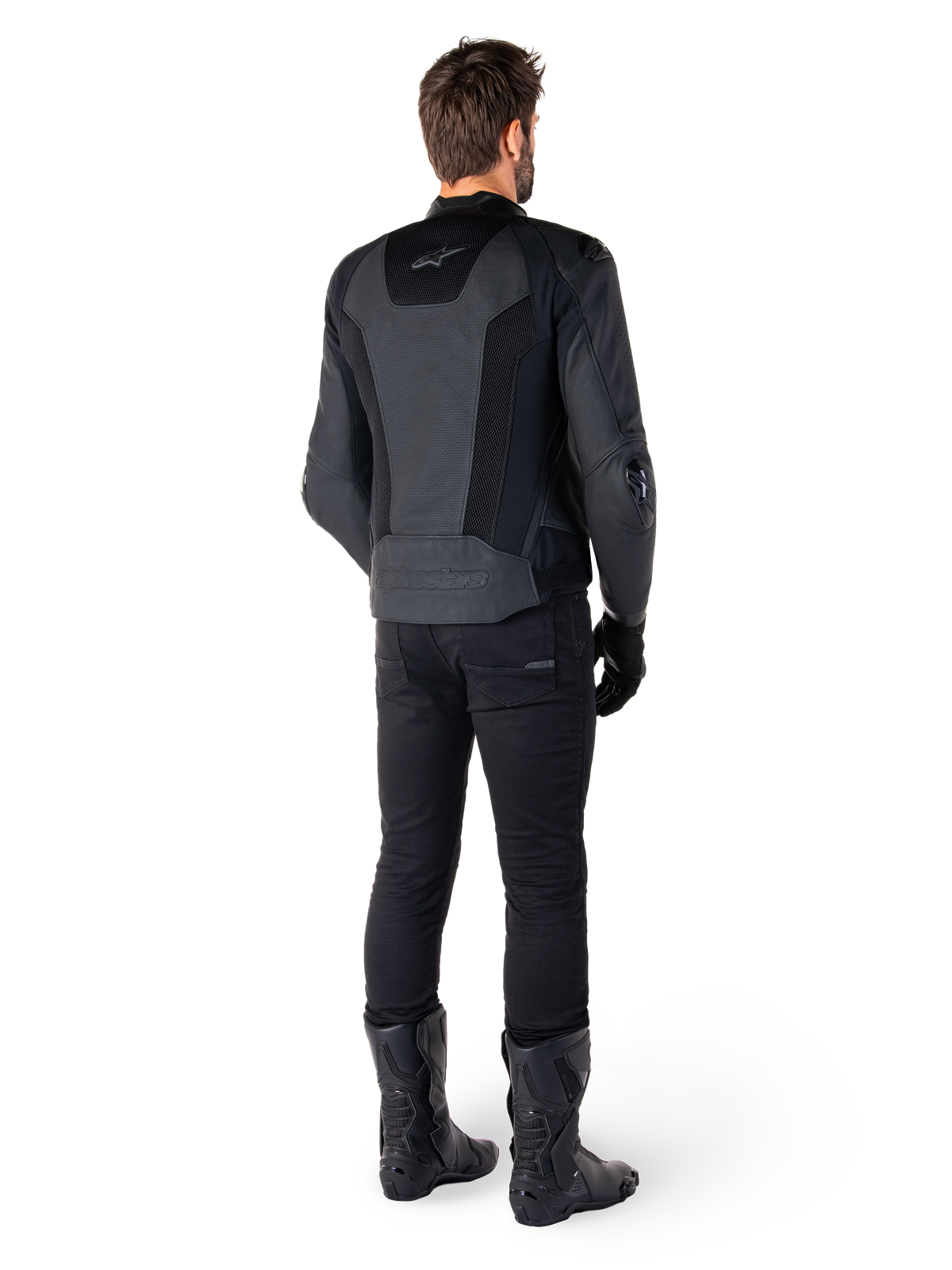 Vista trasera de un hombre con la chaqueta de cuero Alpinestars Missile V3 Airflow en color negro, que presenta amplios paneles perforados para la ventilación, combinada con pantalones y botas de montar negros, mostrando una estética de carreras aerodinámica.