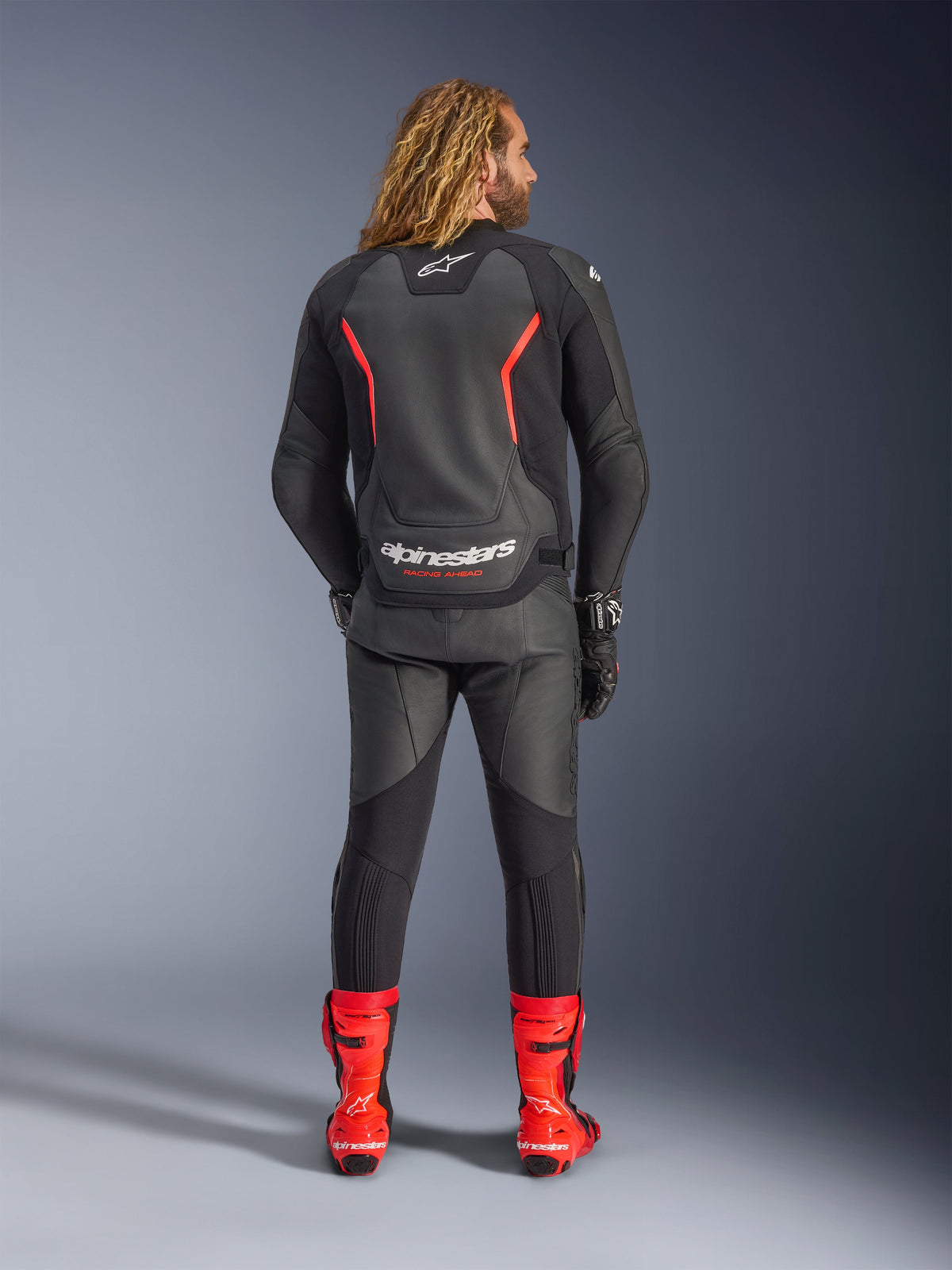 Chaqueta de cuero Alpinestars GP Force V2, negro y rojo flúor, vista desde atrás, combinada con pantalones de cuero negros y botas de carreras rojo brillante, con líneas de acento rojas y logotipo de Alpinestars en blanco en la espalda, equipamiento profesional para motociclismo deportivo