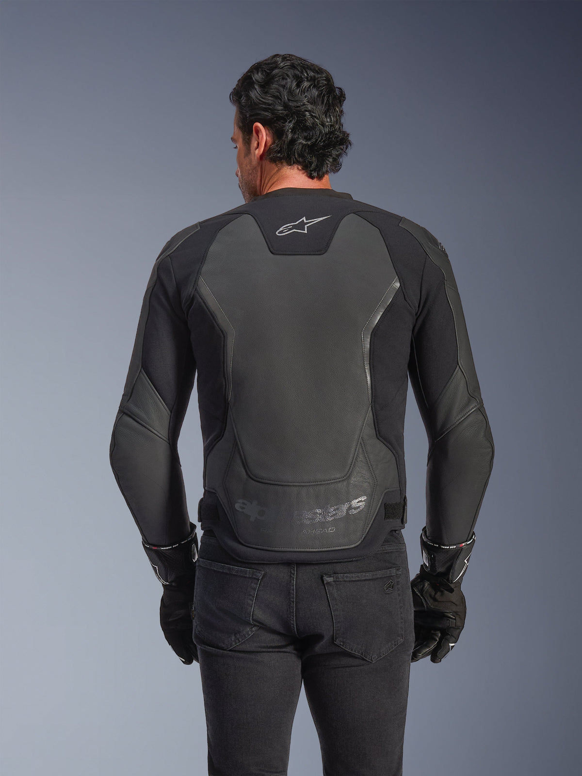 Chaqueta de cuero Alpinestars GP Force V2, negra, vista trasera de un hombre usando la chaqueta con guantes negros y vaqueros, con grandes paneles protectores de cuero en la espalda, logo de Alpinestars en la parte superior trasera y marca grabada en la parte inferior de la cintura, equipamiento de motociclismo de inspiración deportiva