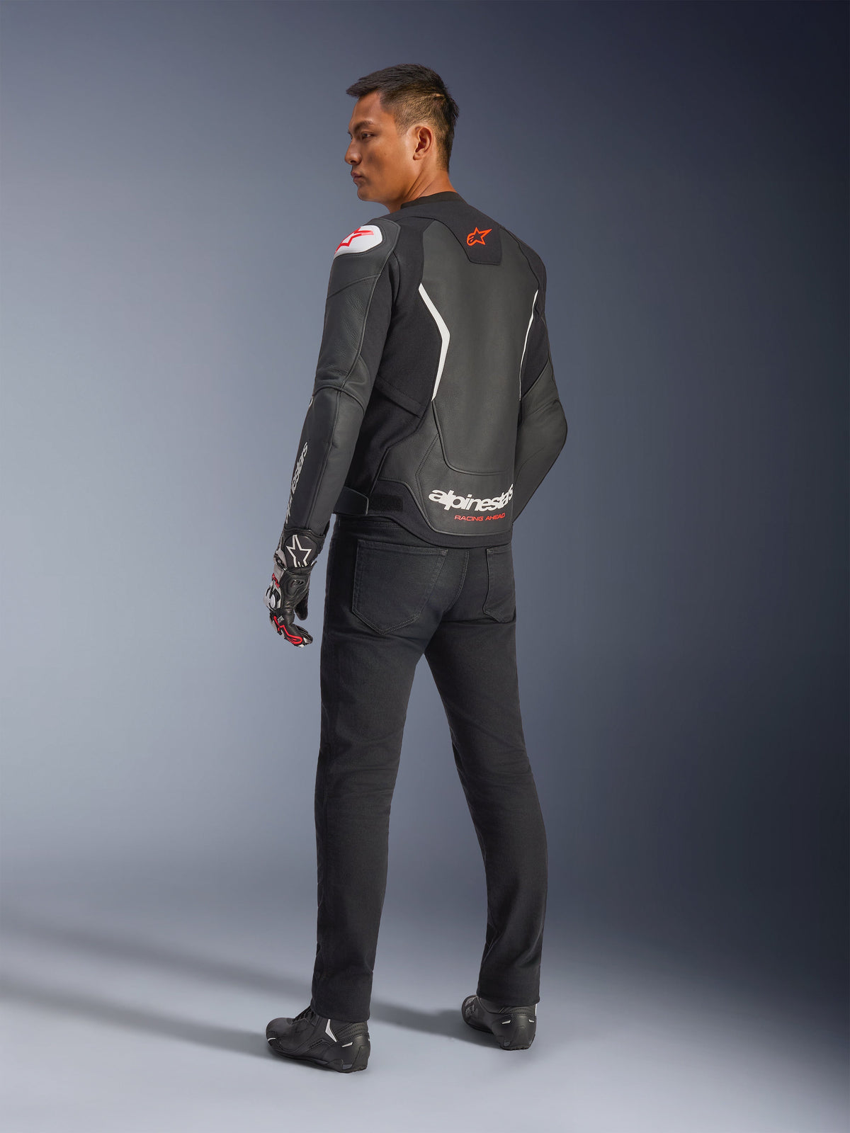 Chaqueta de cuero Alpinestars GP Force V2, negro y blanco, vista posterior que muestra la construcción de cuero con detalles blancos y logotipos rojos, usada con pantalones negros y guantes de carreras, con la marca Alpinestars destacada en la parte baja de la espalda, equipamiento de moto de ajuste deportivo