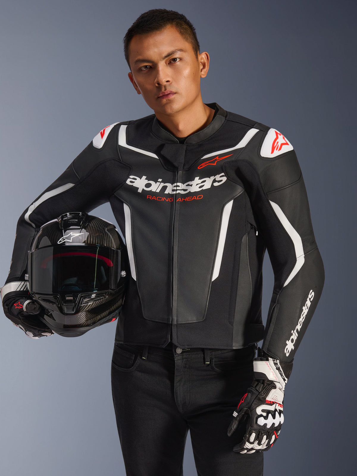 Alpinestars GP Force V2 Leather Jacket, negra y blanca con detalles en rojo, usada por un modelo que sostiene un casco de moto de fibra de carbono y lleva guantes de carreras, con paneles blancos en contraste y el logotipo de Alpinestars.