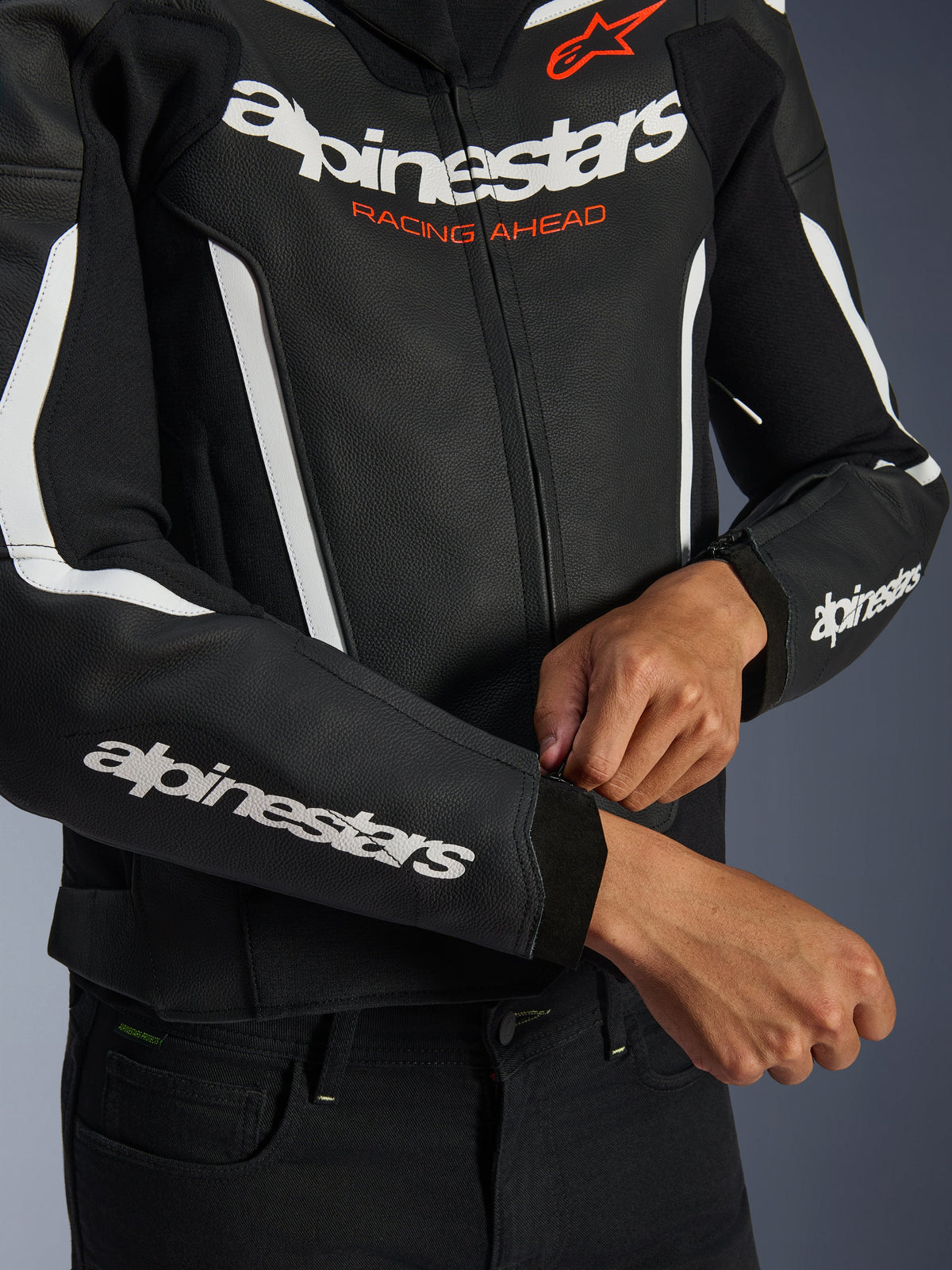 Chaqueta de cuero Alpinestars GP Force V2, negro y blanco, primer plano de un piloto ajustando la cremallera del puño, logotipo blanco en el pecho y las mangas con el texto naranja 'Racing Ahead', cuero bovino de primera calidad para conducción de motos deportivas