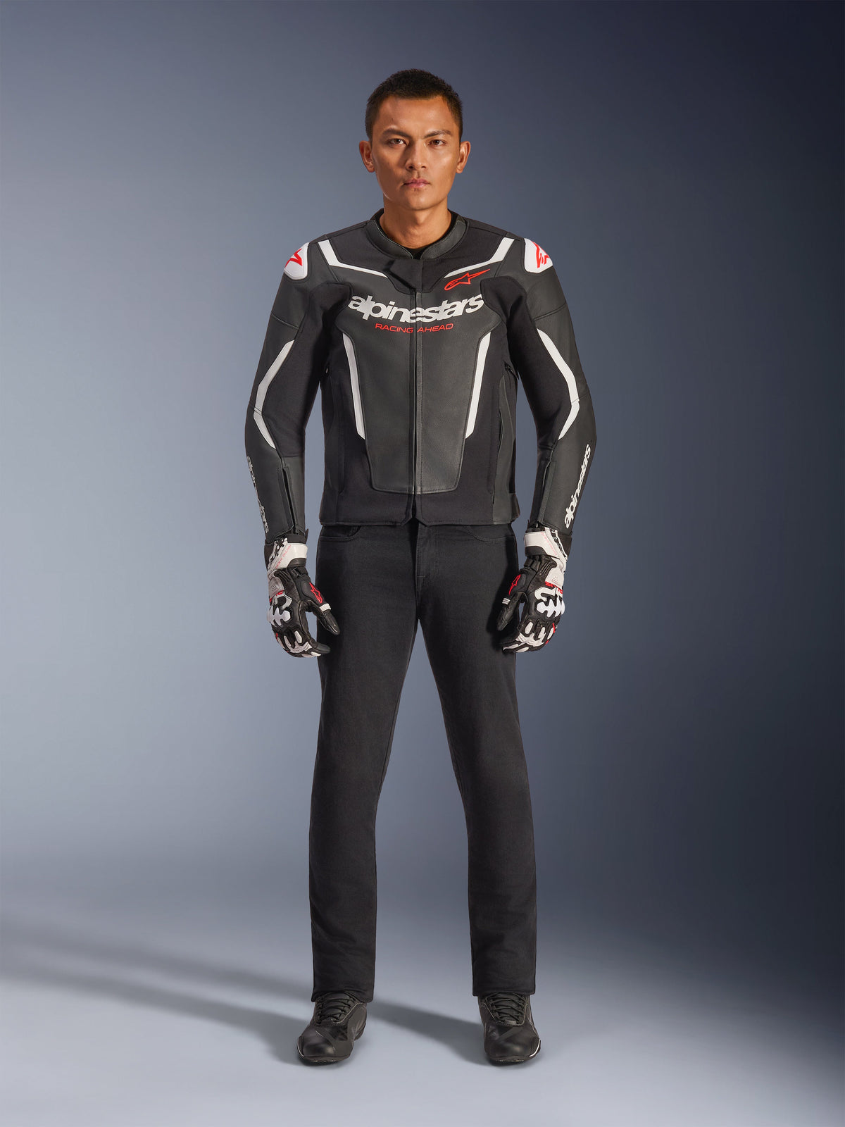 Modelo masculino vistiendo la chaqueta de cuero Alpinestars GP Force V2 en negro y blanco, combinada con pantalones de moto negros y guantes blancos y negros, con logotipos destacados de Alpinestars y detalles rojos en los hombros, equipamiento profesional para conducción deportiva de motocicletas