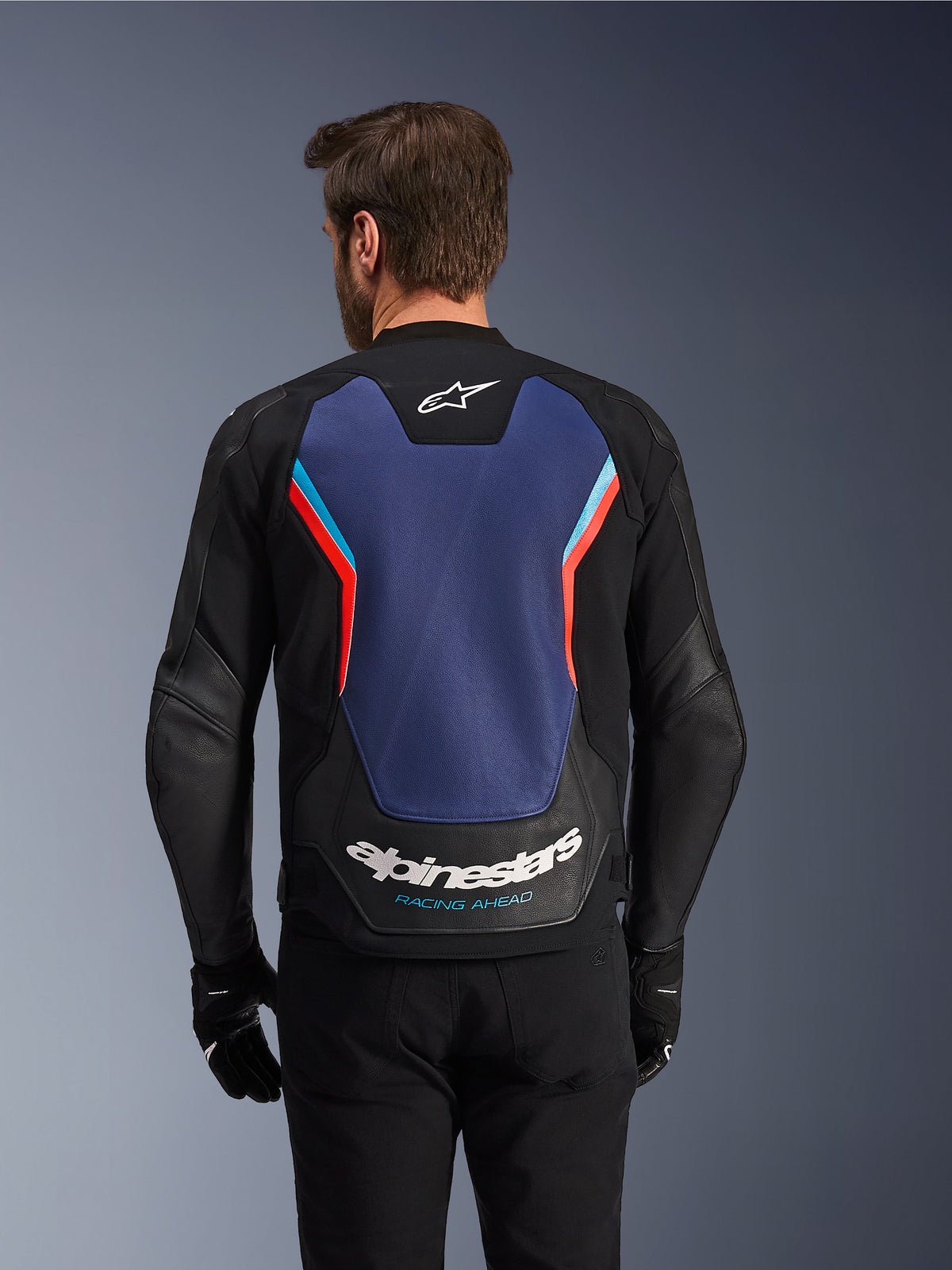 Vista trasera de la chaqueta de cuero Alpinestars GP Force V2 en negro, azul y rojo flúor, con un gran panel trasero azul con detalles en rojo fluorescente y azul claro, logotipo de Alpinestars y marca 'Racing Ahead' en la parte inferior de la espalda, usada por un piloto con guantes y pantalones negros.