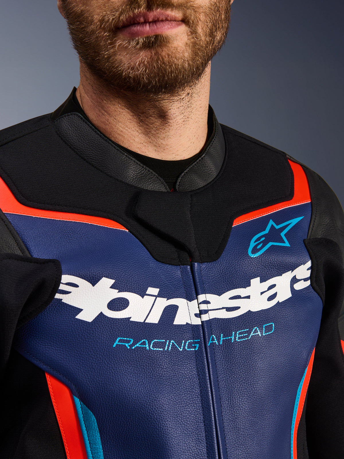 Chaqueta de cuero Alpinestars GP Force V2, vista detallada del pecho y el cuello, azul, negro y rojo flúor, con el logotipo blanco de Alpinestars y el texto 'RACING AHEAD' en azul claro, textura de cuero premium con detalles fluorescentes