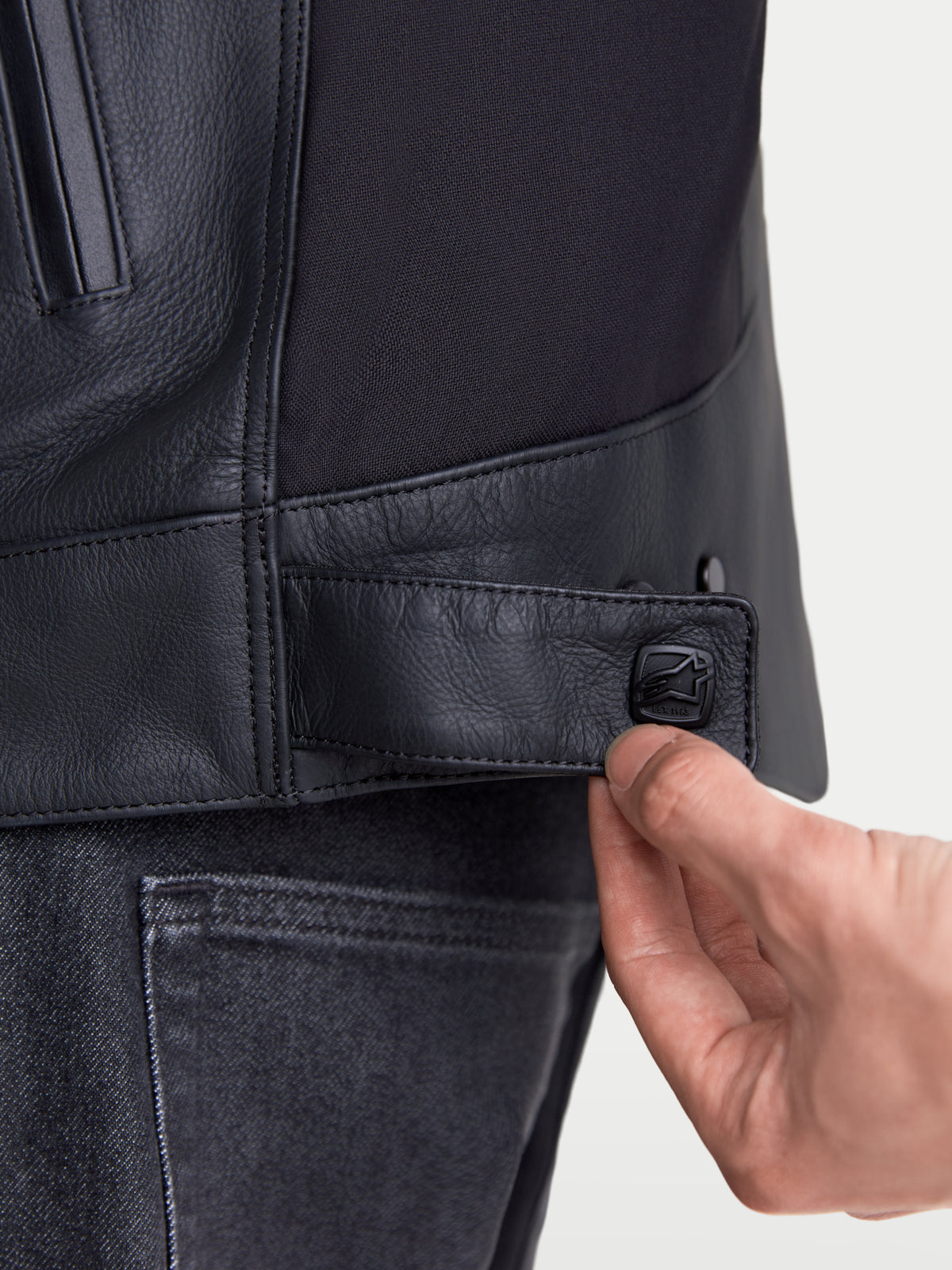 Alpinestars Maver X, chaqueta de moto de cuero urbana para hombre, vista en primer plano de la lengüeta de ajuste con botón a presión en la cintura en color negro, confección en cuero premium