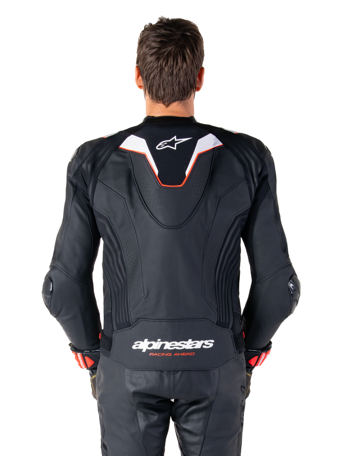 Vista posterior de un piloto con la chaqueta de cuero Alpinestars Missile V3 Ignition, de cuero negro con detalles en blanco y rojo, con una joroba aerodinámica deportiva delgada con logo blanco y la marca 'alpinestars RACING AHEAD' en la parte inferior de la espalda, combinada con pantalones de carreras y guantes de cuero negro a juego.