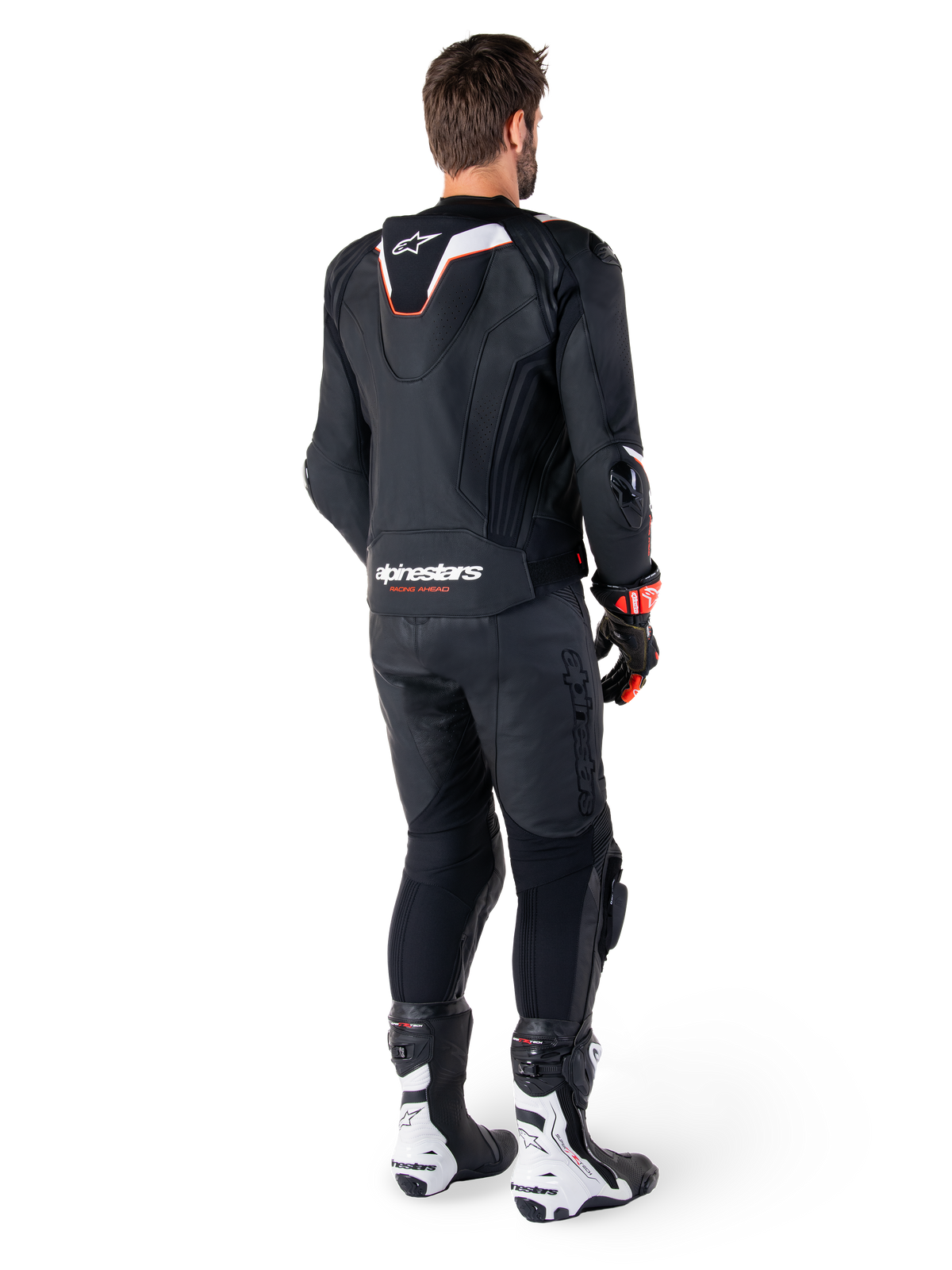 Vista trasera de un piloto con la chaqueta de cuero Alpinestars Missile V3 Ignition en color negro, que presenta una joroba aerodinámica deportiva delgada con detalles en blanco y rojo, logotipo de Alpinestars en la parte inferior de la espalda, combinada con pantalones de cuero negros a juego y botas de competición en blanco y negro; equipamiento profesional de conducción deportiva.