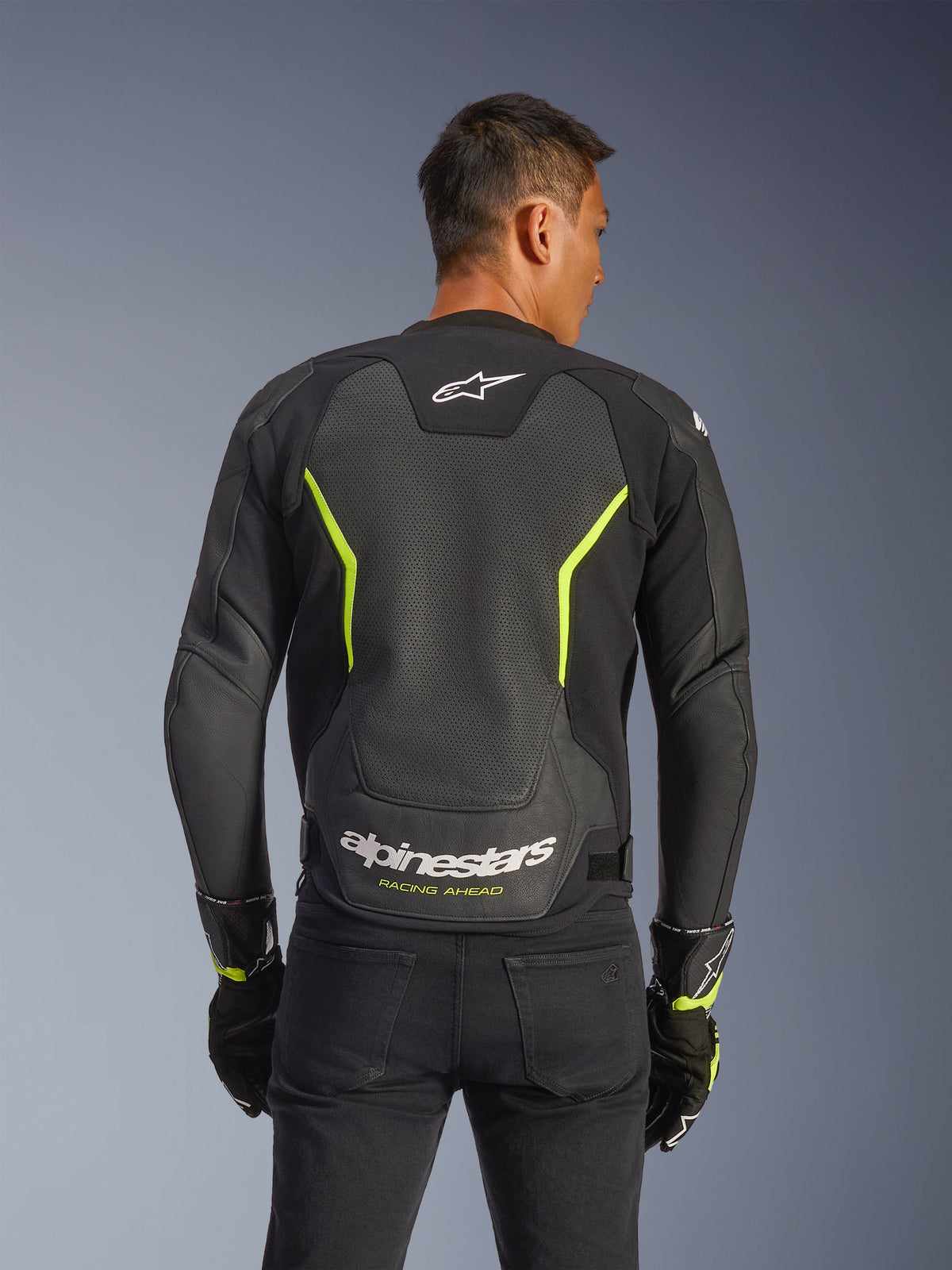 Chaqueta de cuero Alpinestars GP Force V2 Air, negro y amarillo flúor, vista trasera de un piloto masculino que muestra extensos paneles de cuero perforado para el flujo de aire, marca Alpinestars en blanco y texto "Racing Ahead" en amarillo en la parte inferior de la espalda, equipamiento deportivo de moto para conducción en carretera.
