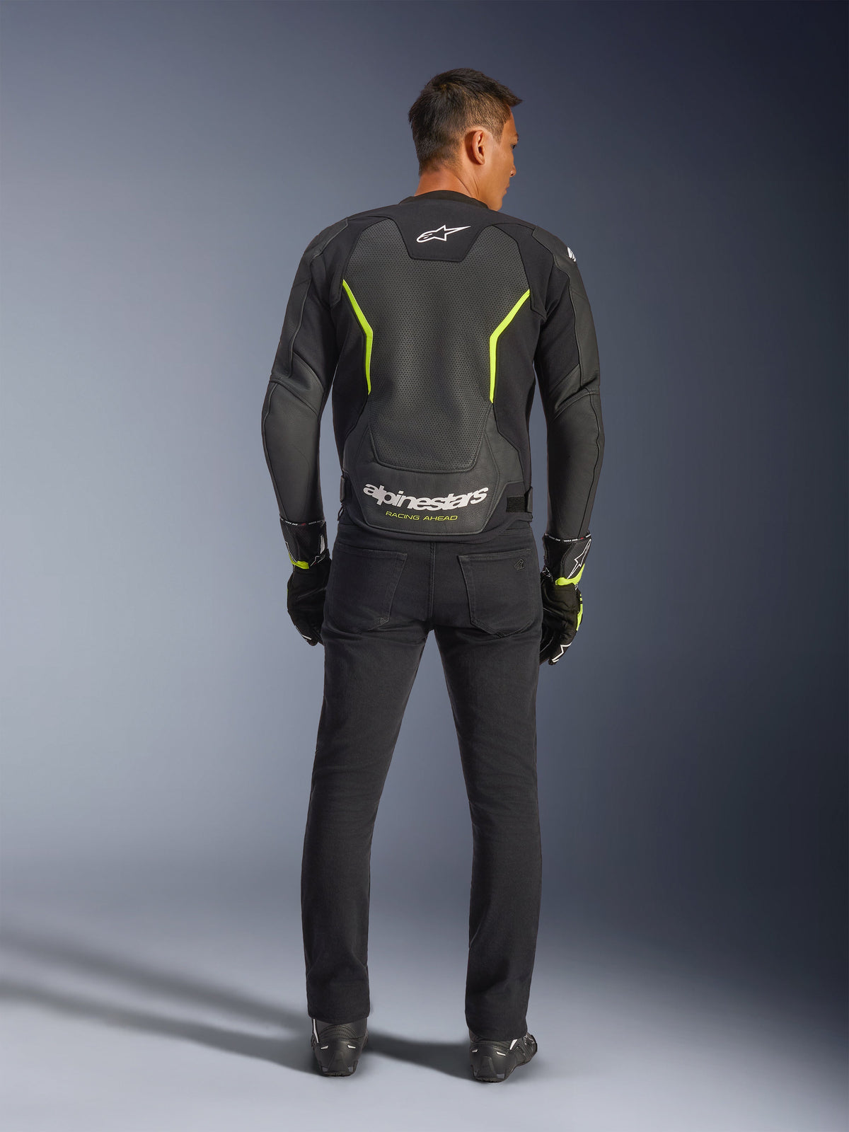 Chaqueta de cuero Alpinestars GP Force V2 Air, negro y amarillo flúor, vista posterior de modelo usando la chaqueta deportiva de moto, con amplias secciones perforadas, detalles en amarillo neón y logotipo blanco de 'alpinestars' en la parte baja de la espalda, combinada con equipamiento de conducción negro