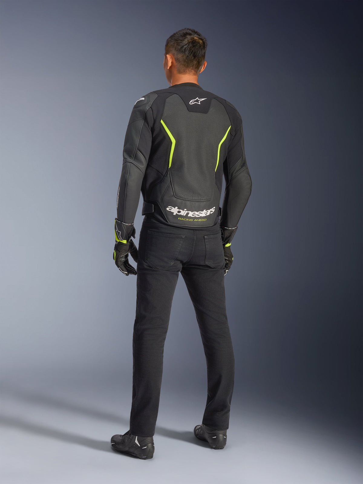 Chaqueta de cuero Alpinestars GP Force V2 Air, negro y amarillo flúor, vista trasera de un motorista usando la chaqueta de cuero perforado, con detalles amarillos angulares y logotipo blanco de Alpinestars con 'RACING AHEAD' en la parte baja de la espalda, combinada con pantalones y botas de motorista negros.