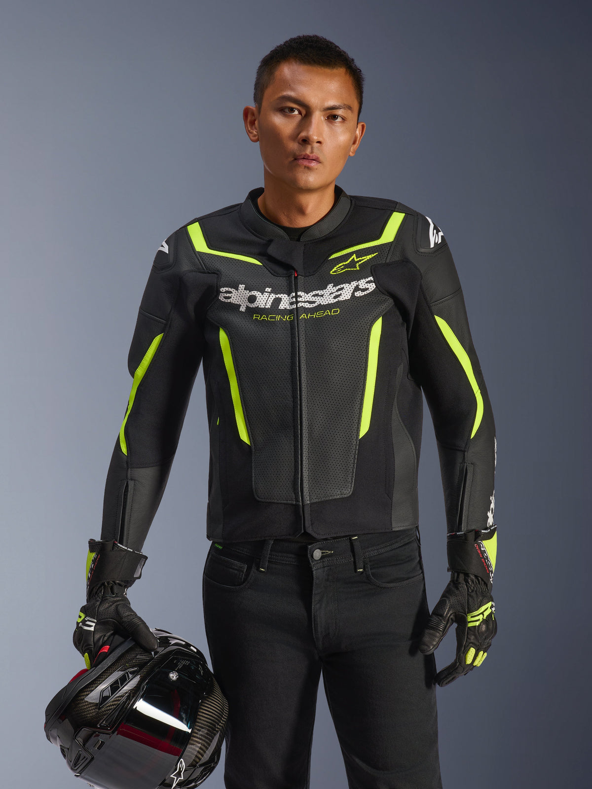 Piloto masculino con la chaqueta de cuero Alpinestars GP Force V2 Air en negro y amarillo flúor, sujetando un casco de moto negro, con cuero perforado, logotipos de Alpinestars en blanco y detalles en amarillo neón, combinada con guantes de carreras a juego y pantalones negros