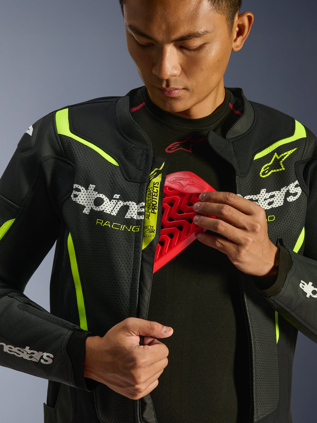 Chaqueta de cuero Alpinestars GP Force V2 Air, negro y amarillo flúor, chaqueta de moto de cuero perforado, mostrada por un piloto que enseña el compartimento del pecho con un inserto protector rojo, con logotipos blancos de Alpinestars y amplias perforaciones para el flujo de aire.