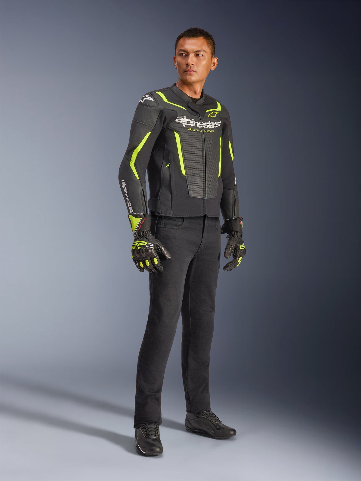 Modelo masculino luciendo la chaqueta de cuero Alpinestars GP Force V2 Air en negro y amarillo flúor, combinada con pantalones de conducción negros, guantes a juego y calzado negro; fotografía de estudio de cuerpo completo de equipamiento deportivo para moto con cuero perforado y logotipos llamativos.