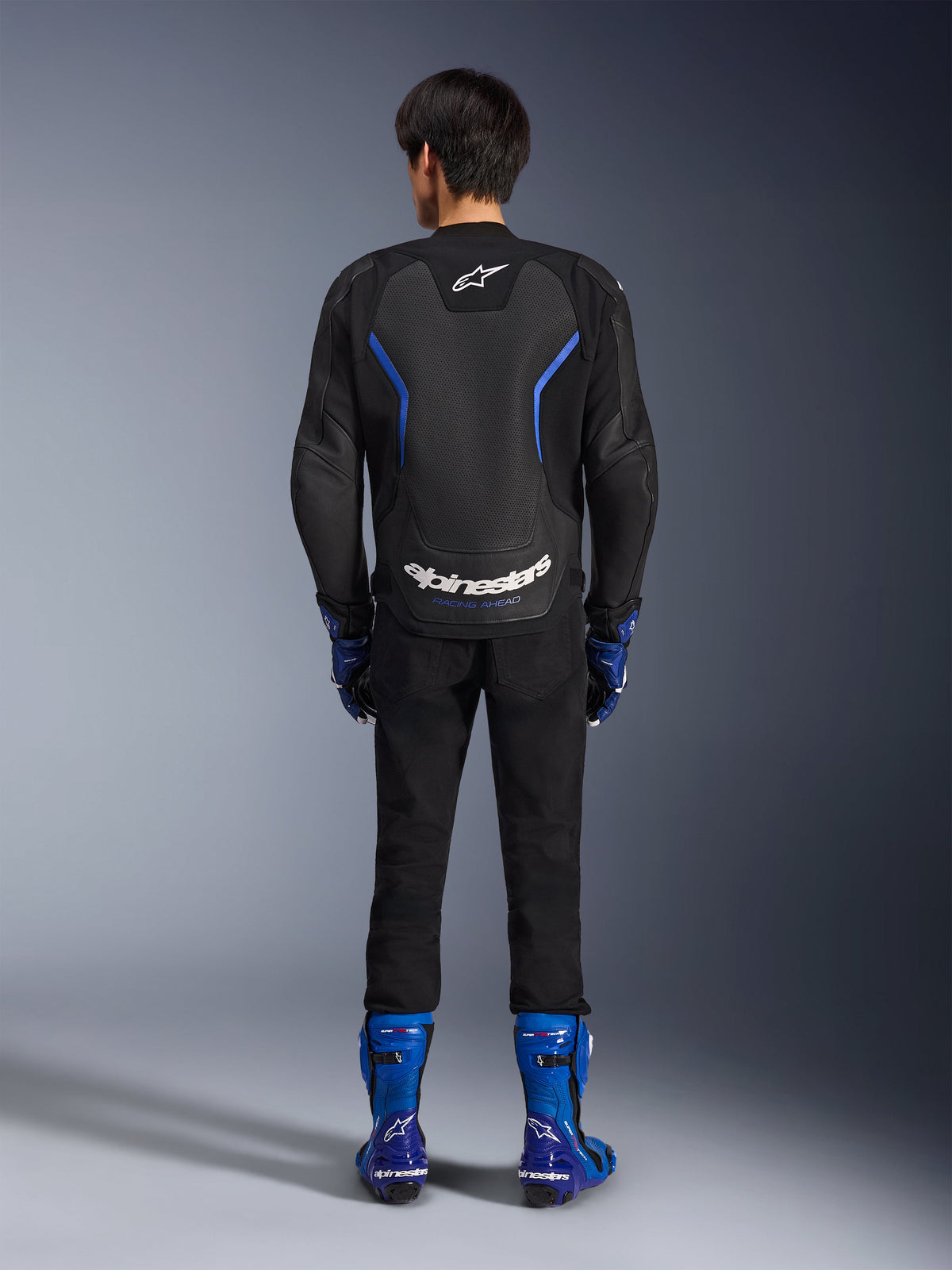 Chaqueta de cuero Alpinestars GP Force V2 Airflow, negro y azul metálico, vista posterior que muestra los paneles de cuero perforado y la marca en blanco, combinada con botas de carreras azules y pantalones negros, equipamiento de motociclismo deportivo