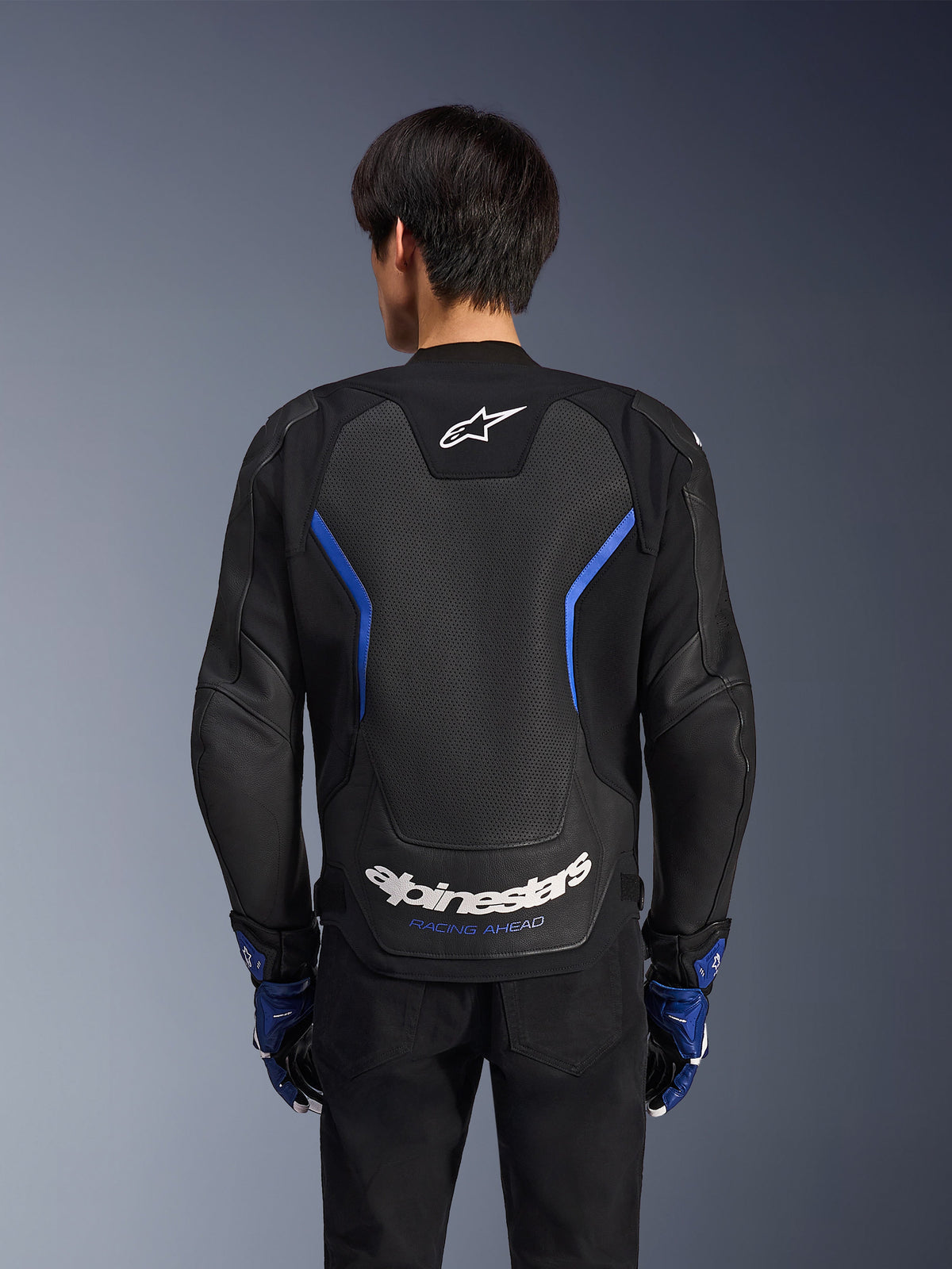 Chaqueta de cuero Alpinestars GP Force V2 Airflow, Negro Azul Metálico, vista trasera, con un gran panel trasero perforado para ventilación, detalles en azul y logotipos de Alpinestars en blanco, usada con guantes azules a juego y pantalones negros para conducción deportiva.