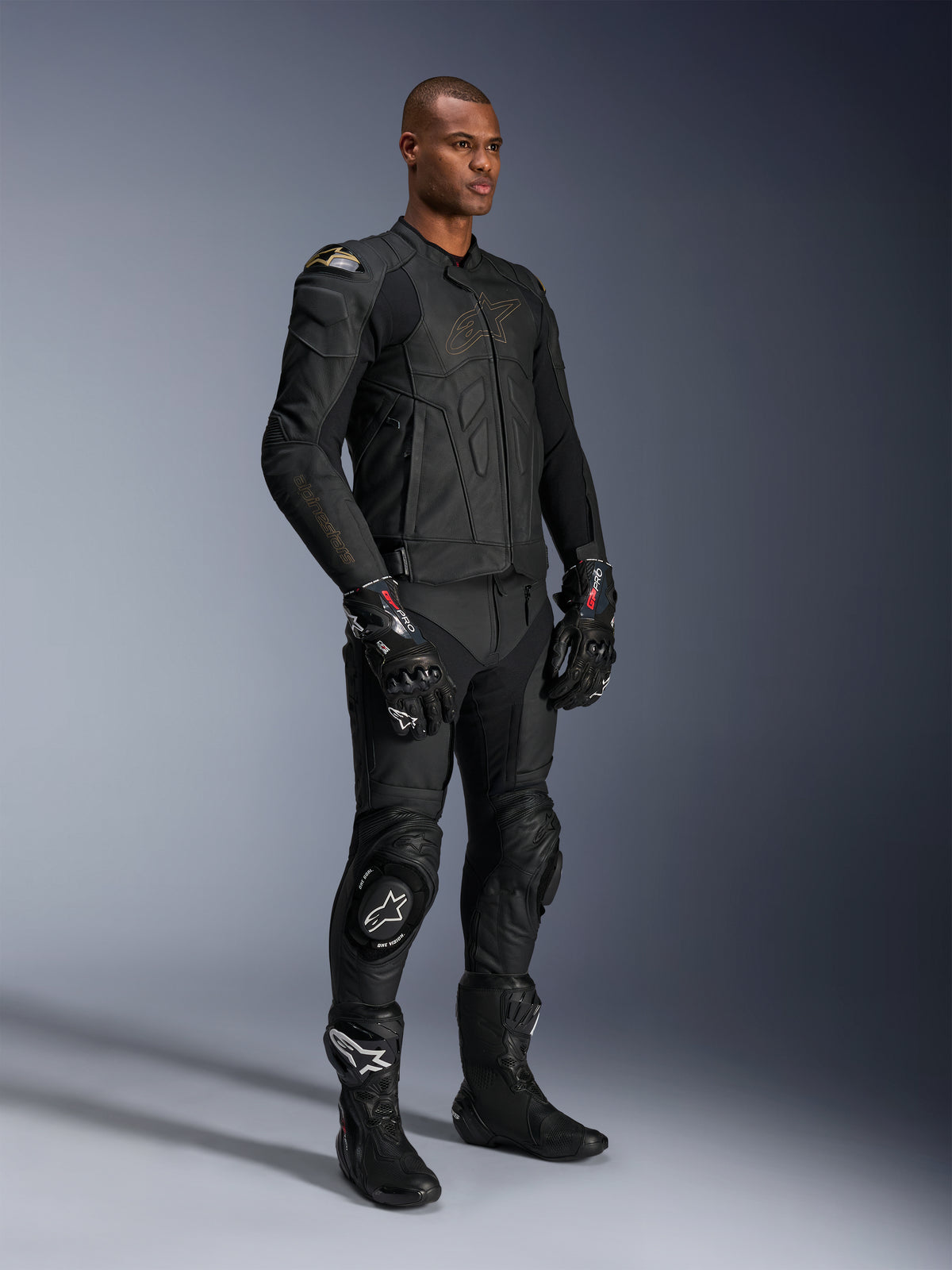 Modelo masculino con pantalones de cuero Alpinestars Missile V3 y chaqueta de cuero negra a juego con detalles dorados, guantes de competición GP Pro negros y botas de carreras negras, equipo completo de motociclismo para carretera, foto de estudio